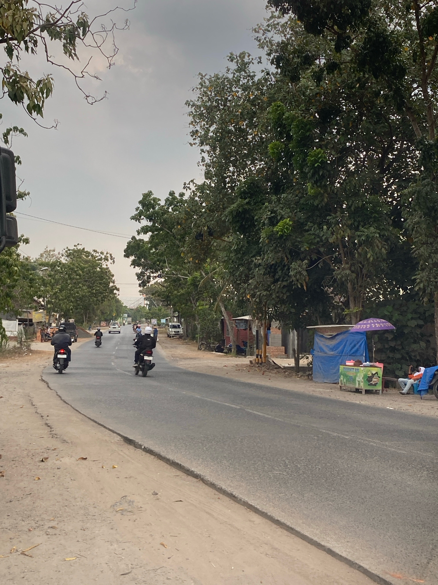The image captures a street scene in Metro, Indonesia, under an overcast sky. A paved road runs through the frame, with a dusty shoulder on the left. Several motorcycles are in motion on the road. In the foreground, two motorcyclists are heading away from the viewer, their bodies cloaked in dark attire. Further down the road, more motorcycles and a white car are visible.
The right side of the road is lined with trees and small structures. A roadside stall with a blue tarp roof and a green display case is present, topped with a purple umbrella. A person in an orange shirt sits on a bench next to the stall. Another person on a motorcycle is parked nearby. Buildings and more trees line the background, with distant hills or mountains faintly visible. The overall impression is of a typical day on a road outside the main urban center.