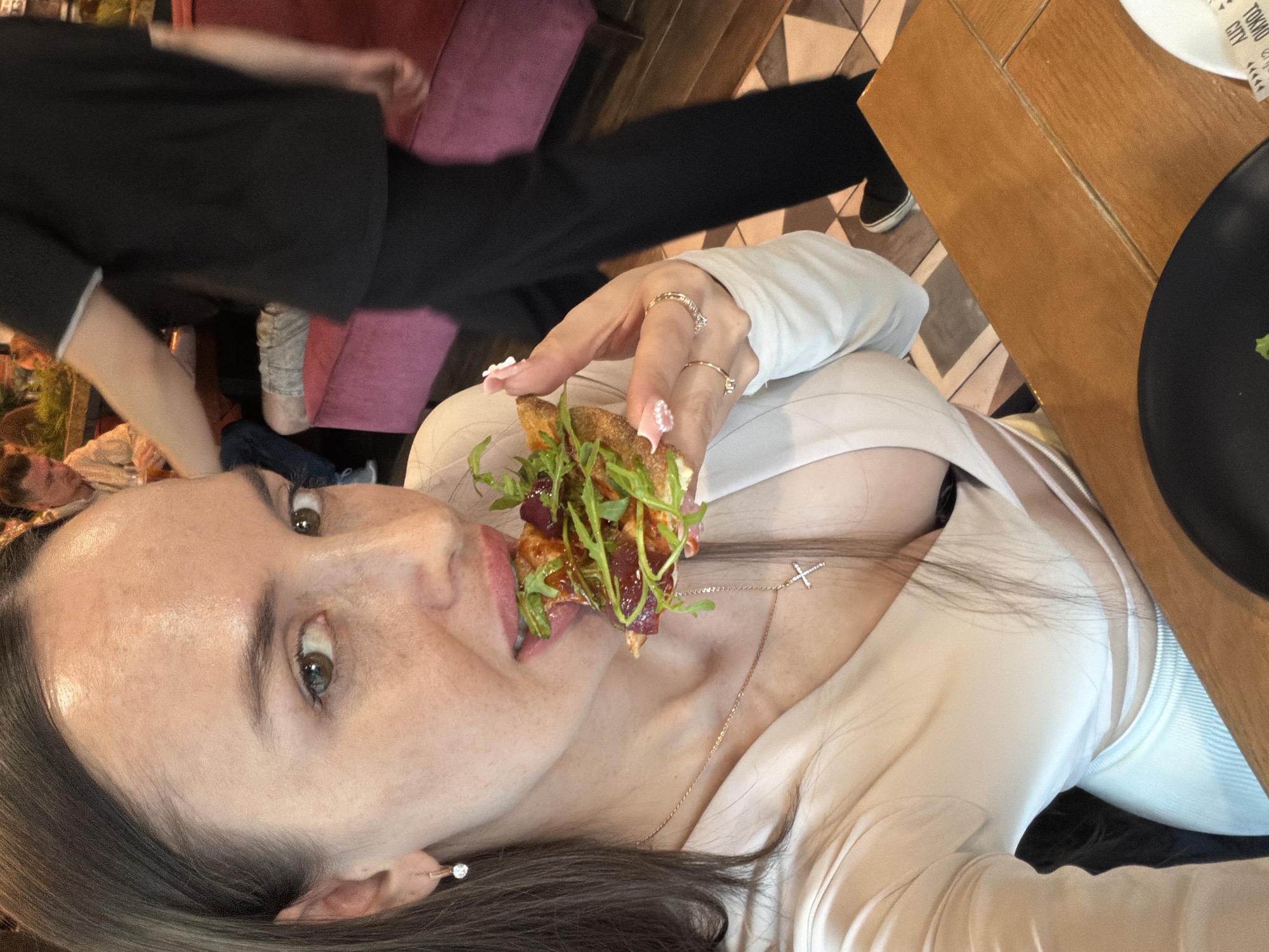 The image depicts a woman seated at a wooden table in an indoor restaurant or cafe, specifically a Tokyo City establishment in Saint Petersburg, Russia.
The central subject, a woman estimated to be between 25 and 35 years old, occupies the foreground. She has long, dark brown hair, a visible part on her left side, and light brown eyes. Freckles are discernible on her nose and cheeks. She is wearing a light cream-colored, long-sleeved top with a deep V-neck, revealing a black bra. A light blue ribbed garment is visible below the table line. Her right earlobe displays a single, round, clear-stoned stud earring. A delicate rose gold-toned chain necklace with a small, clear-stoned cross pendant hangs around her neck. Her right hand, exhibiting manicured fingernails with a pink and white gradient design, holds a slice of food. This food item, resembling a flatbread or pizza, is topped with a red sauce, thinly sliced dark red meat, and a generous quantity of green arugula, along with some smaller dark red vegetable pieces. The woman is in the act of biting into this food. She wears three gold-toned rings on her right hand: a thin band on her ring finger, a textured band on her middle finger, and a simple band on her pinky finger.
In the mid-ground, to the left, a man with light brown hair and a beard, wearing a beige collared shirt, is seated at a table. To his right, an individual in a black short-sleeved polo shirt with white trim and black trousers is in motion, walking from left to right.
The environment includes a light-colored wooden table where the woman is seated. A black plate is partially visible on the table to the bottom left, and a white paper napkin with "TOKYO CITY" printed on it in black is visible to the bottom right. Seating in the background consists of a magenta-colored upholstered sofa or banquette. The floor features a geometric tile pattern in various shades of brown and beige. Some green foliage is visible on a surface in the background behind the seated man.