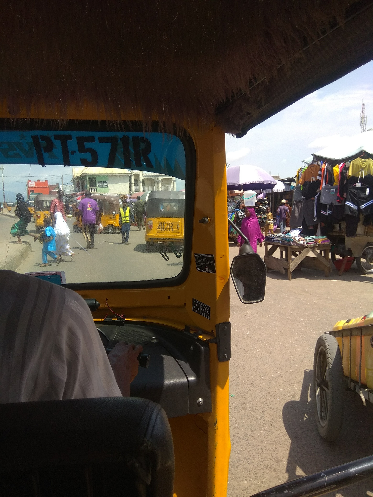 The image captures a street scene in Jalingo, Nigeria, as viewed from the interior of a yellow auto-rickshaw. The rickshaw's dashboard and a portion of the driver's shoulder and arm are visible in the foreground, with the driver's hand on the steering wheel. Through the windshield, a bustling street unfolds. Several other yellow auto-rickshaws are present, some occupied by passengers. People are seen walking and crossing the street. One auto-rickshaw has the license plate "4RZR". On the right side of the street, market stalls display clothing, with vendors tending to their goods. A white and purple umbrella shades part of the market. The sky is bright and appears to be a clear day, with a distant telecommunications tower visible. The road surface is asphalt. The overall impression is of a busy urban environment, typical of a market day in a Nigerian city.