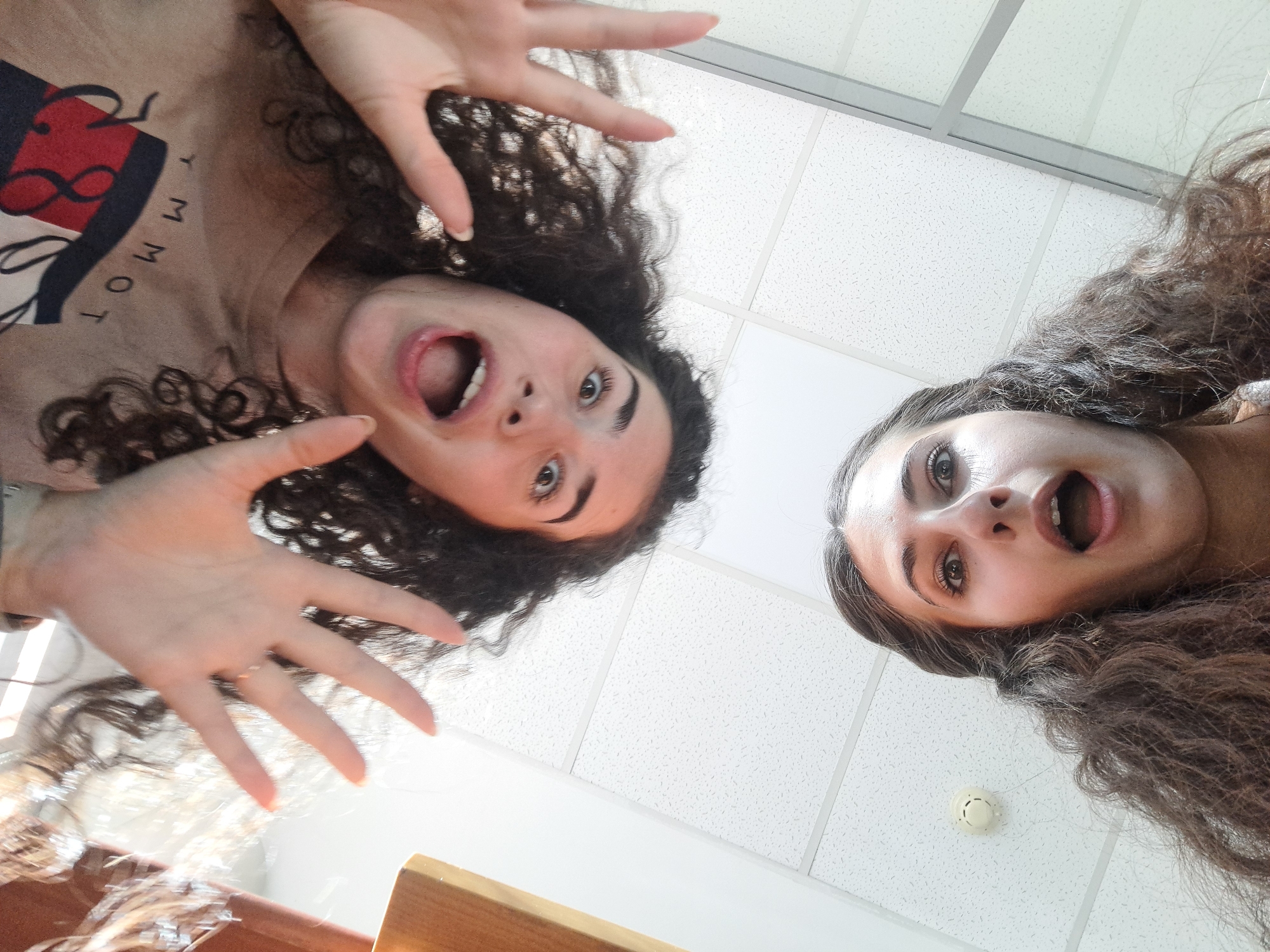 Two young women with curly hair are angled downward towards the camera. One woman on the left wears a Tommy Hilfiger shirt and has her hand raised towards the camera. Both women have their mouths open in surprise. The background shows a white tile ceiling, a smoke detector, and what appears to be a wooden surface. The location is Unknown, Unknown.