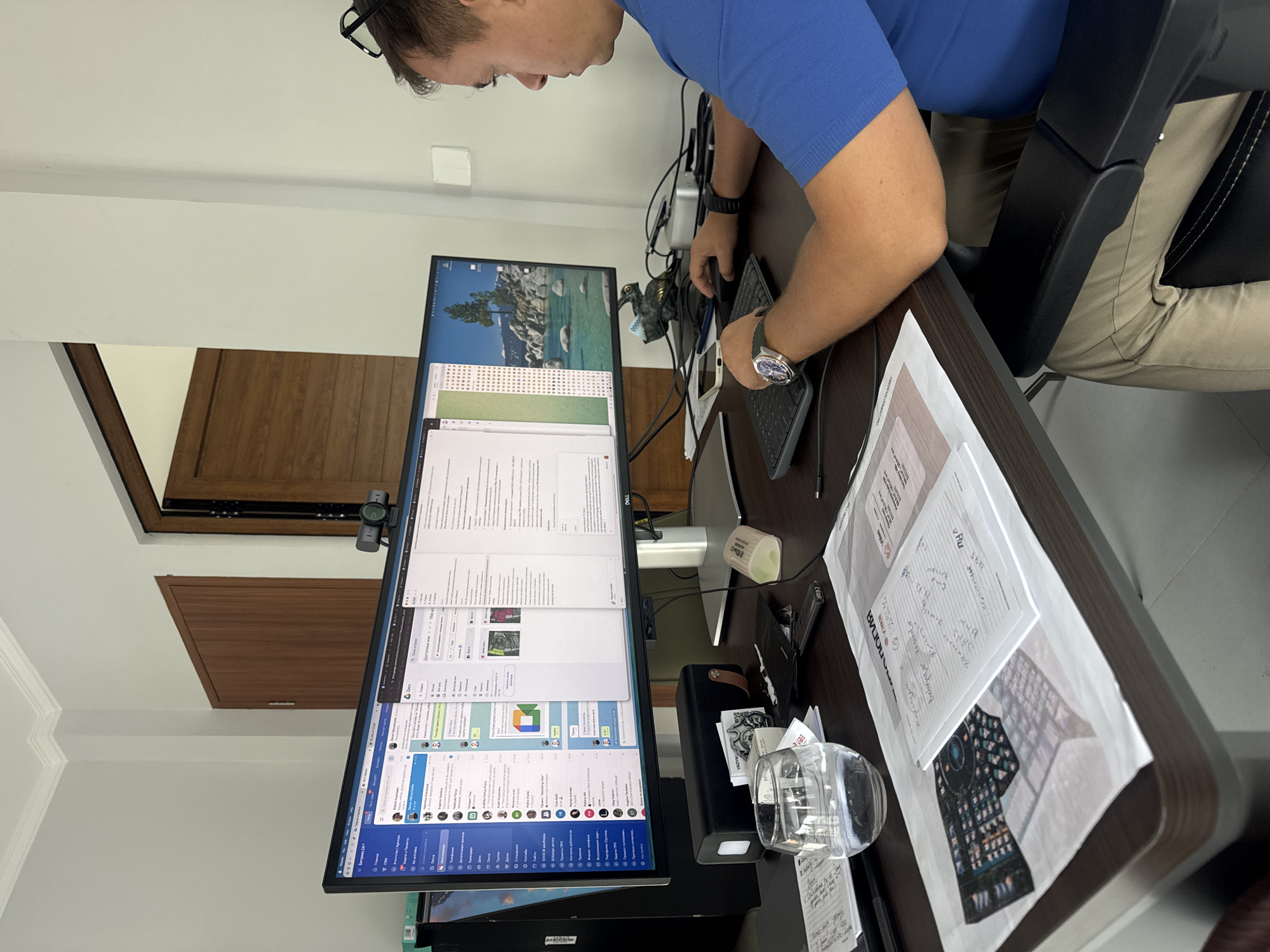 An overhead, slightly angled view captures a man working diligently at a computer setup in what appears to be an office or home workspace in Denpasar, Indonesia. The man, wearing a blue short-sleeved shirt, khaki pants, and a wristwatch, is seated in a dark office chair, leaning forward and typing on a black keyboard with his right hand while his left hand rests near a mouse. Glasses are pushed up onto his head.
His workstation features two large monitors, vertically stacked, displaying various open applications. The top monitor shows a scenic desktop background of a lake and mountains, along with a grid-like application. The bottom monitor is filled with multiple windows, including what appear to be chat applications (resembling Telegram and Slack or similar platforms), a document editor, and other software. Much of the visible text on the screens is in Cyrillic, with recognizable terms like "Битрикс24" (Bitrix24) and "Документы" (Documents) visible. A webcam is mounted on top of the bottom monitor.
The desk, made of dark wood, is cluttered with work-related items. These include a power strip with several cables, a small decorative pelican figurine, a white mug, a clear glass, a black portable speaker or power bank, and several sheets of paper. On one paper, handwritten notes and calculations are visible, along with the printed word "ВИТА" (VITA). The background of the room features white walls and a light-colored wooden door or paneling. The lighting suggests daytime. The man appears focused and engrossed in his work.