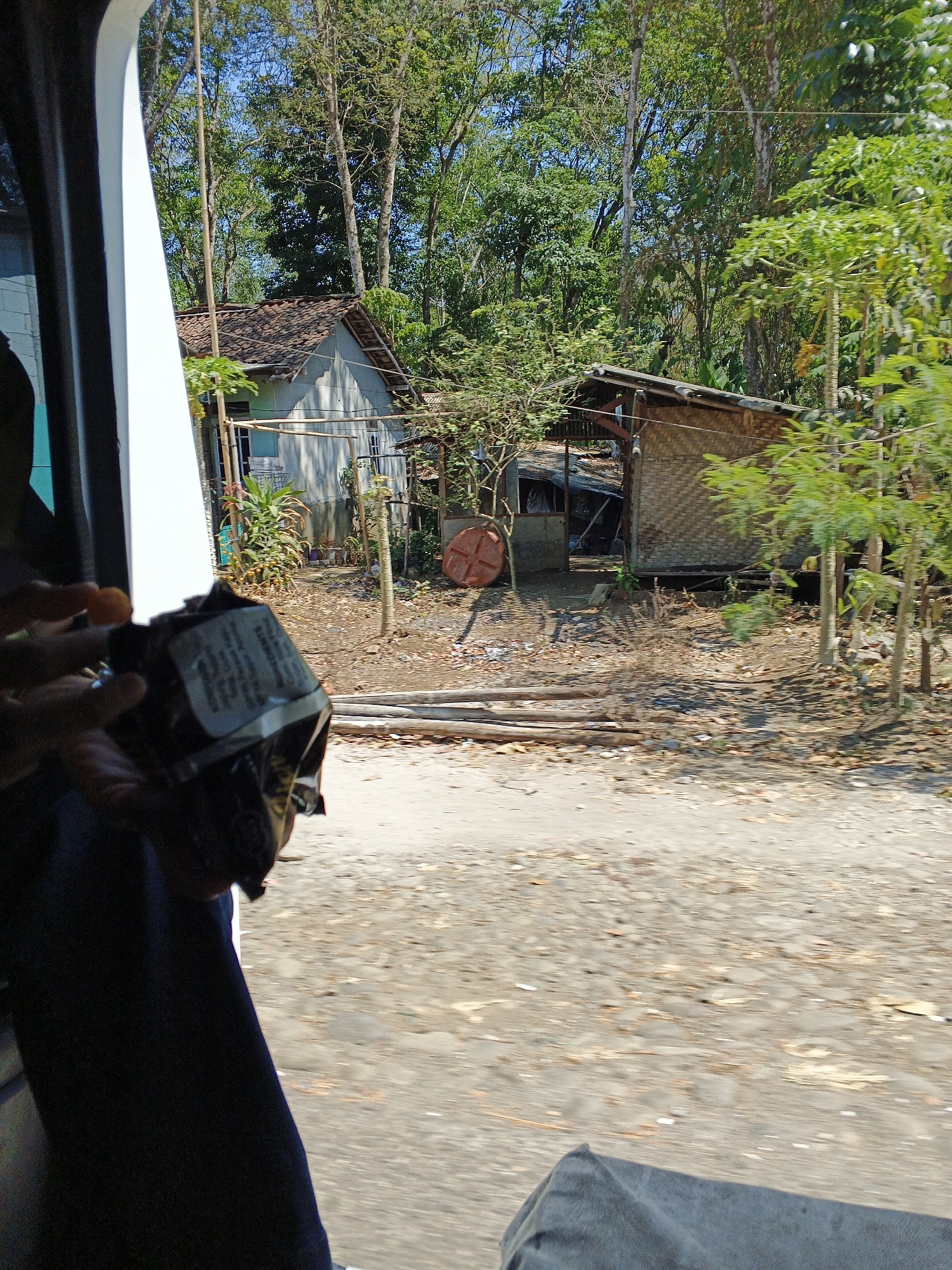 A view from inside a vehicle looking out at a rural scene in Warungkiara, Indonesia. In the foreground, a person's dark blue pants are visible. Their left hand is holding a dark-colored package, possibly food, with some text on it. The package appears partially opened, and a small orange object, possibly food, is held near the package by the hand.
The outdoor environment shows a dirt and gravel road. Beyond the road, there are two rustic structures. The building on the left is a small, light-colored house with a tiled roof. A clothesline is visible in front of it. To the right, there is a smaller, open-sided structure with a corrugated metal roof and walls made of woven bamboo. A round, red object, possibly a drum or container, is situated near this structure.
The background is dominated by lush green trees, suggesting a natural, possibly forested, area. The sky is clear and blue, indicating sunny weather. The overall impression is of a simple, rural dwelling surrounded by nature. The motion blur in the image suggests the photo was taken while the vehicle was in motion.