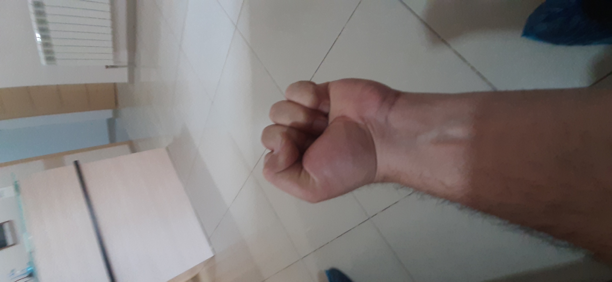 The image shows a close-up of a clenched fist and forearm. The subject's skin is light-toned and appears to have some light hair growth on the forearm. The fist is tightly closed, with knuckles prominently visible. The background is out of focus but suggests an indoor environment with tiled flooring and what appears to be furniture and a radiator in the distance. There is no identifiable information linking this image to Yessentuki, Russia.