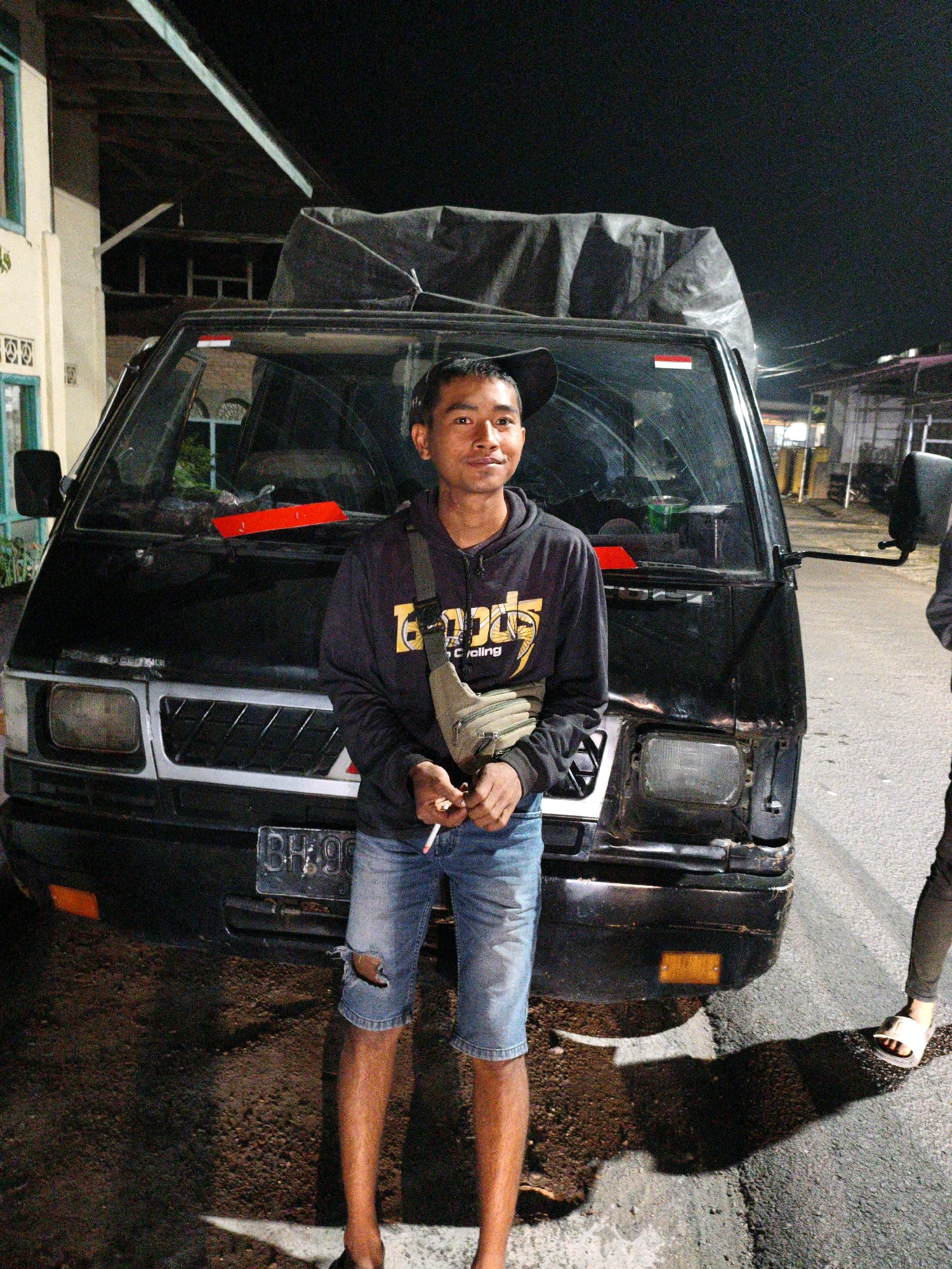 A young man stands outdoors at night in front of a black van. He is wearing a dark hooded sweatshirt with yellow lettering, denim shorts with a tear on the left thigh, and a light-colored fanny pack across his chest. He is holding two unlit cigarettes in his right hand. The van has a tarp covering its roof and appears to be parked on a wet asphalt road. The license plate is partially visible and reads "BH 90". The background shows a building on the left and other structures in the distance, illuminated by artificial lighting. The scene suggests a location in a city or town, potentially in Koto Baru Semurup, Indonesia, given the context.