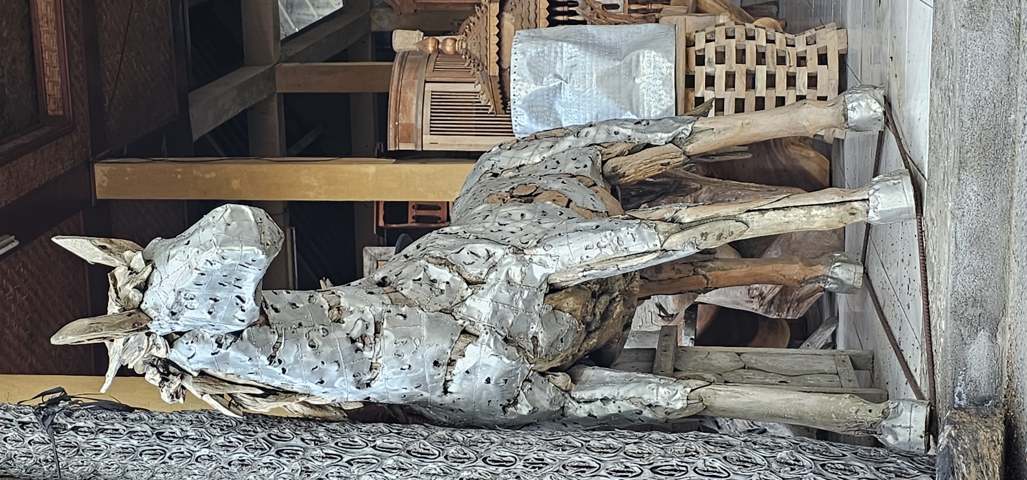 The image shows a sculpture of a horse made from wood and metal scraps, lying on a patterned surface. The horse is composed of wooden branches and logs, covered with patched sheets of silver-colored metal. The metal has numerous perforations and irregular cuts. The legs are primarily wooden, and are capped with a wider band of the same silver metal. The background is filled with wooden objects and architectural elements, creating a cluttered setting. The scene is located in Ubud District, Indonesia.
