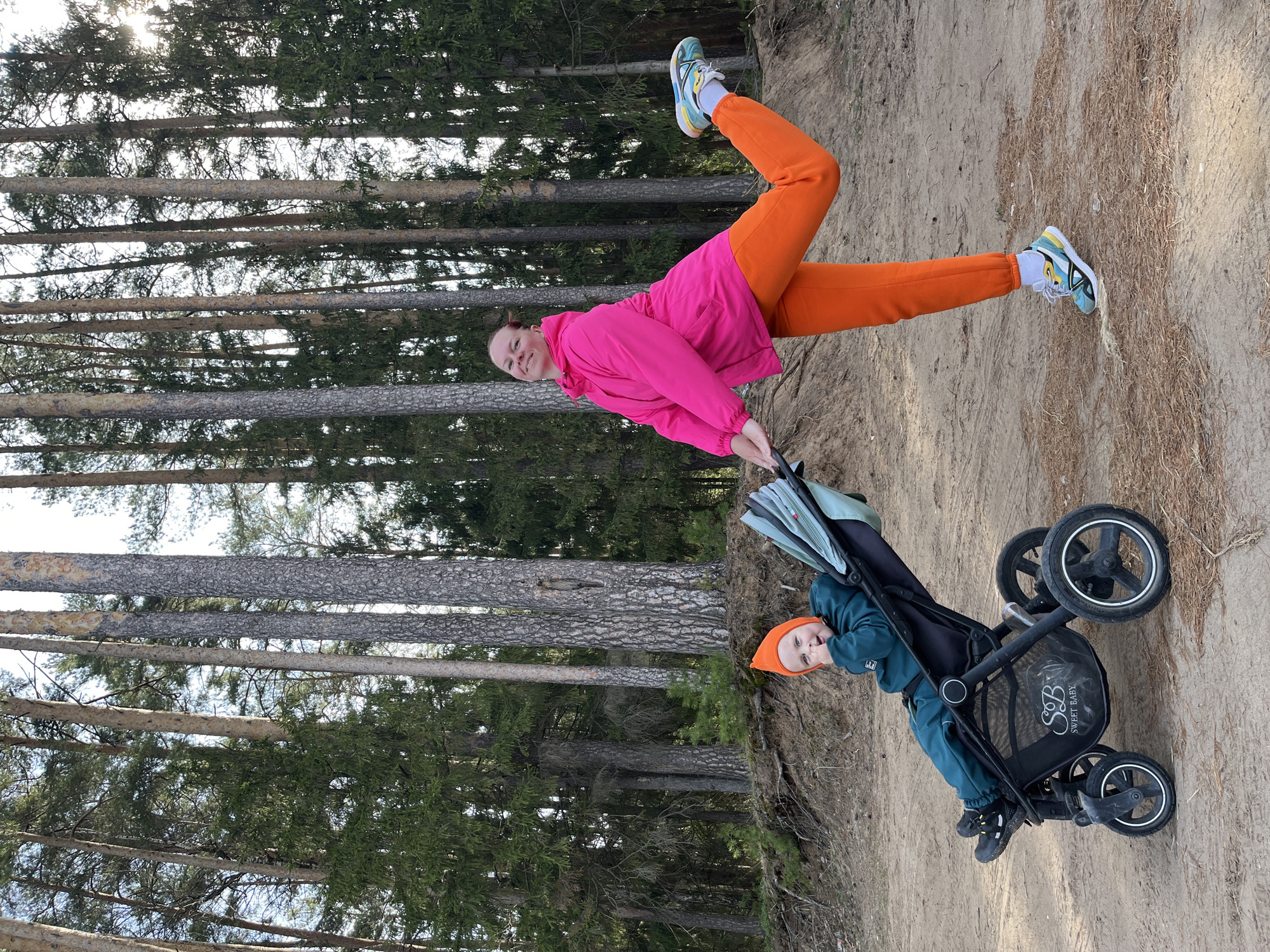 A woman and child in a stroller are positioned in front of a wooded area. The woman, wearing a bright pink jacket and orange pants, is standing on a sandy path. She holds the stroller, in which a child wearing an orange hat and teal clothing sits. The wooded area consists of tall, slender trees. The scene is lit by sunlight. The location is Chaikovsky, Russia.
