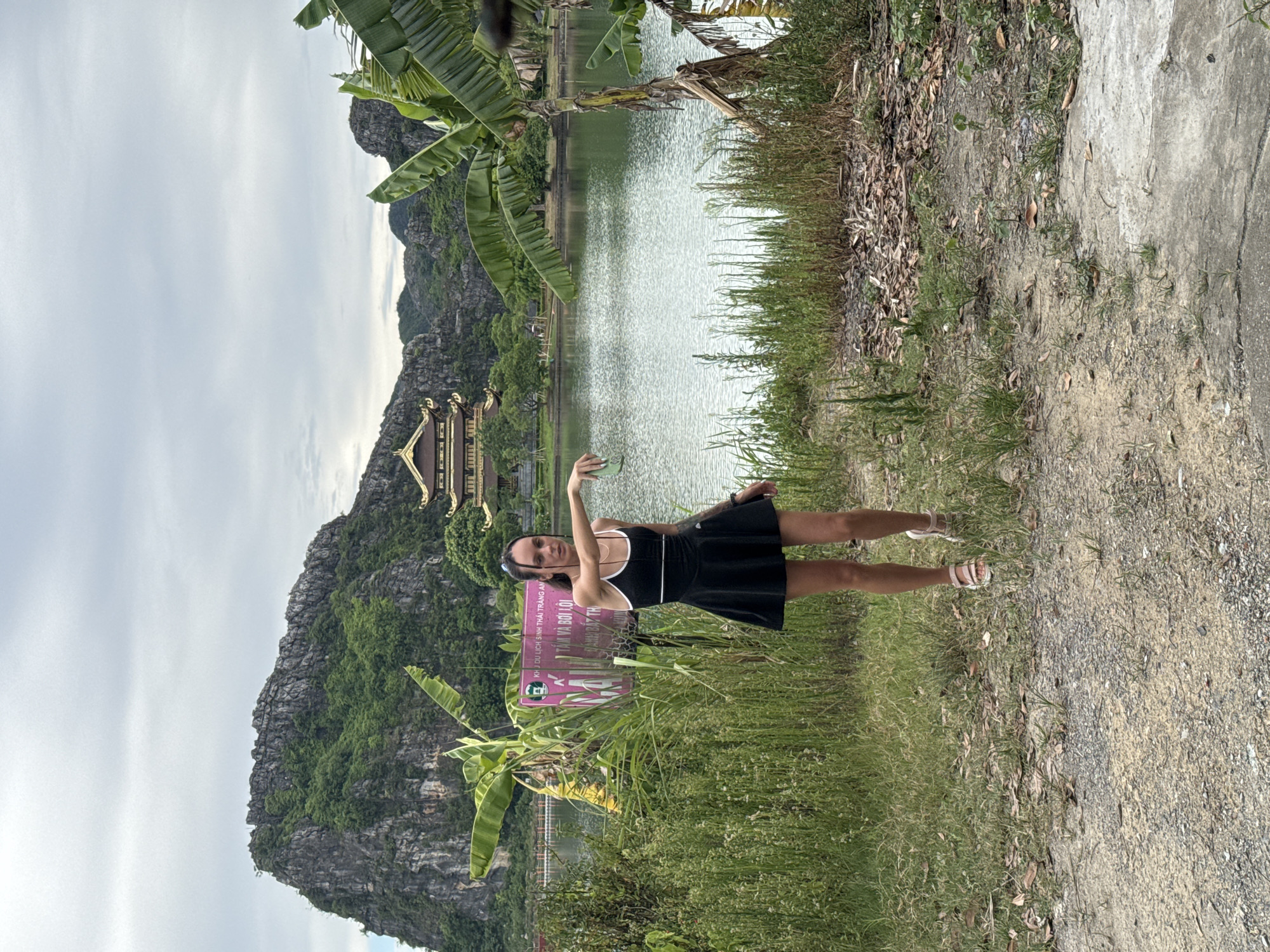 A woman stands in front of a body of water with a pagoda-style building and karst mountains in the background. The woman is wearing a black dress with white trim and pink sandals. She has her left arm extended, holding a green object. The environment is lush and green, with tall grasses and banana trees. A signpost with Vietnamese text is partially visible to the left of the woman. The sky is overcast. This scene is likely located in Hoa Lư Ward, Vietnam, an area known for its natural beauty and historical sites.