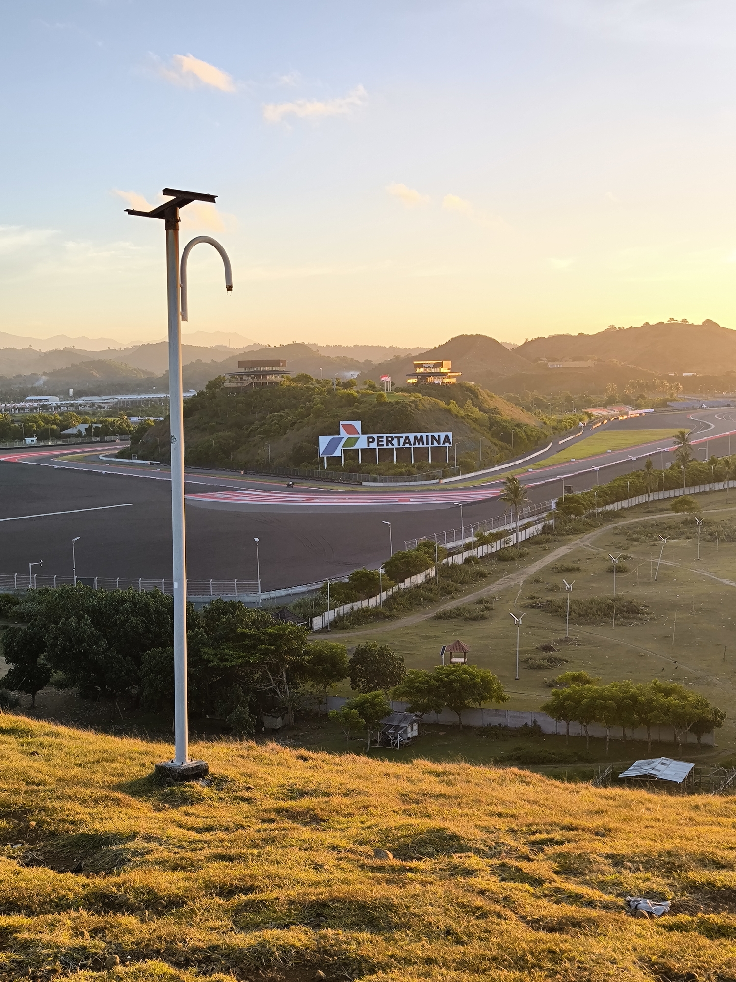 The image depicts the Pertamina Mandalika International Street Circuit in Kuta, Indonesia at sunset. The foreground features a grassy hill with a tall, white metal pole structure. The circuit is visible in the midground, including the asphalt track, red and white rumble strips, and perimeter fencing. A large "PERTAMINA" sign is displayed on the hillside adjacent to the track. Further in the background, the landscape transitions into hills and mountains with sparse vegetation. A multi-story building is visible on top of one of the hills. The sky shows a gradient of colors, from blue near the top to orange and yellow near the horizon, indicating the sunset. The overall lighting suggests a warm, golden hour.