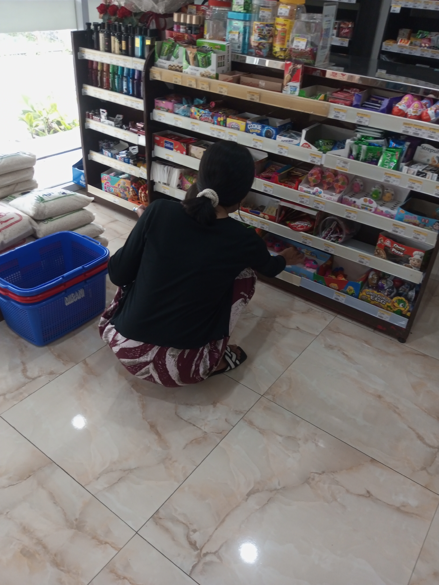 A woman wearing a black shirt and patterned maroon and white pants is kneeling in front of shelves stocked with various goods in a store. She is reaching out to the shelves. In the foreground, there is a blue plastic basket and stacks of white sacks. The floor is tiled with a marble pattern. The store appears to be located in Andongsili, Indonesia.