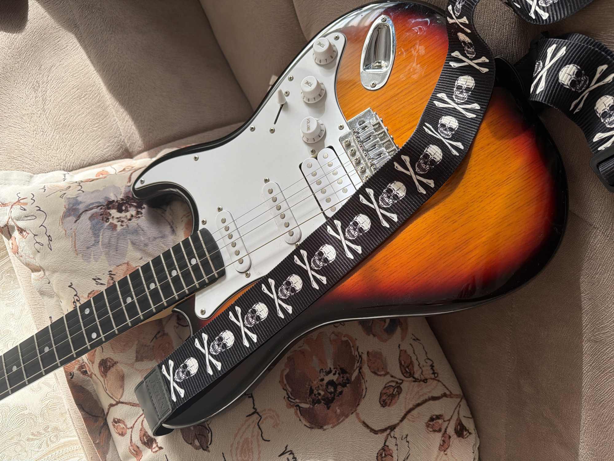 The image shows a close-up, high-angle shot of a sunburst electric guitar with a black strap featuring a skull and crossbones pattern. The guitar is resting on a couch with a patterned cushion. The environment is a domestic interior, likely a living room. There are no people visible in the image. The scene is well-lit, with natural light casting shadows on the couch. There is no visible indication of the location being in Baku City, Azerbaijan.