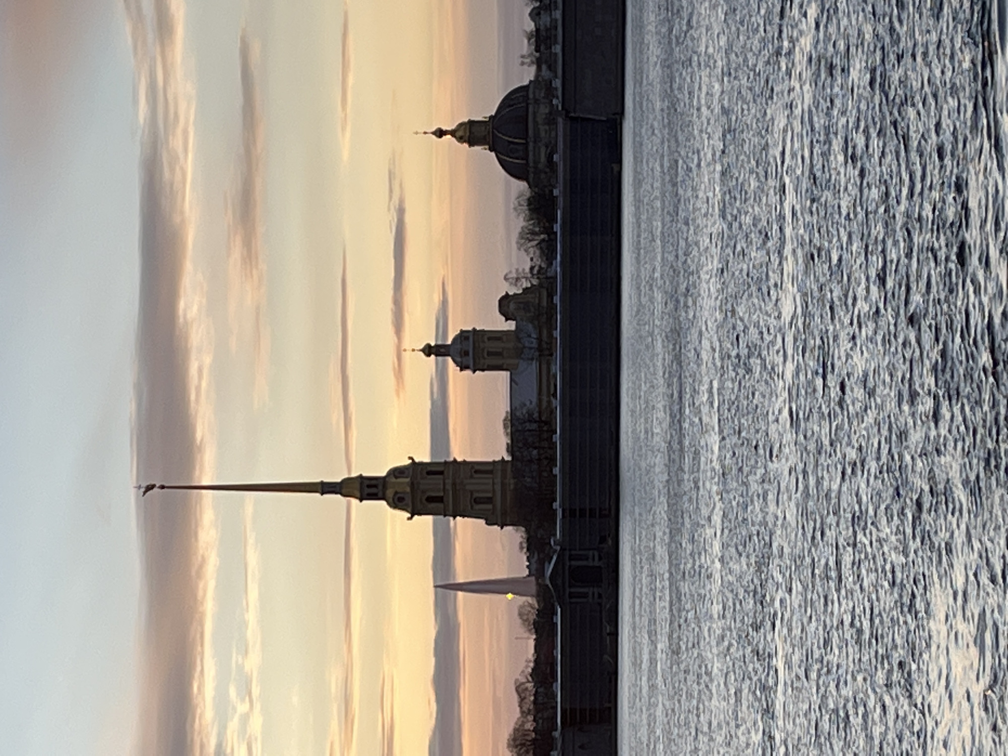 The image depicts a sunset view of the Peter and Paul Fortress in Saint Petersburg, Russia, as seen from across the Neva River. The fortress's iconic golden spire and cathedral dome are visible against a sky with colorful hues of pink, orange, and blue. The Neva River is textured with the movement of water.