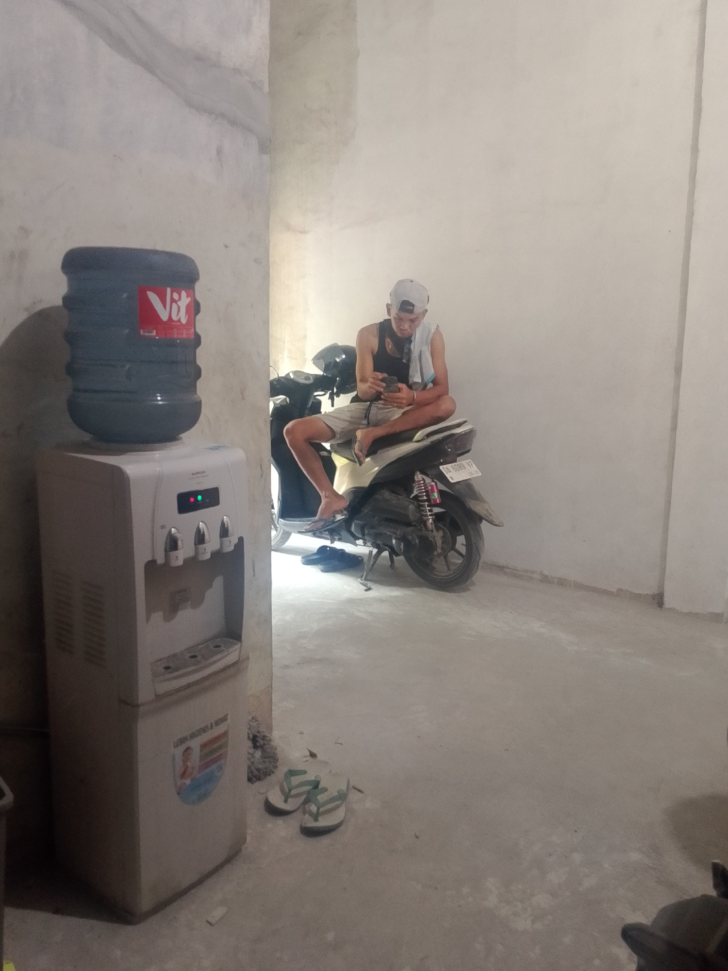 A young man wearing a black tank top, grey shorts, a grey baseball cap, and a light-colored towel around his shoulders is seated on a scooter. He is looking down at his phone. The scooter is parked in what appears to be an unfinished building with rough concrete walls and a grey concrete floor. To the left of the scooter, there is a white water dispenser with a large blue water bottle on top. The water dispenser has three spigots and indicator lights. A pair of flip-flops is visible on the floor near the water dispenser. The scene is dimly lit. This image was taken in Banjarmasin, Indonesia.