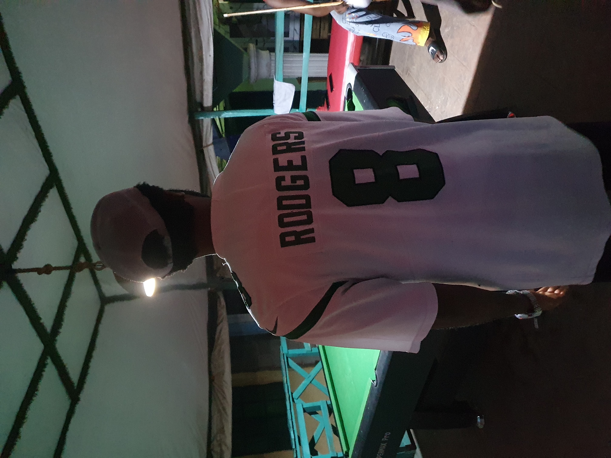 A person in a "RODGERS 8" jersey stands next to a pool table, potentially playing a game of billiards. The location seems to be indoors, likely in a recreational room or under a sheltered area, suggested by the structural elements in the background. The environment is illuminated by overhead lighting. This is a potential residential area or social gathering spot, but there are no indications this scene is located in Lokoja, Nigeria. More information is needed for a city specific location.