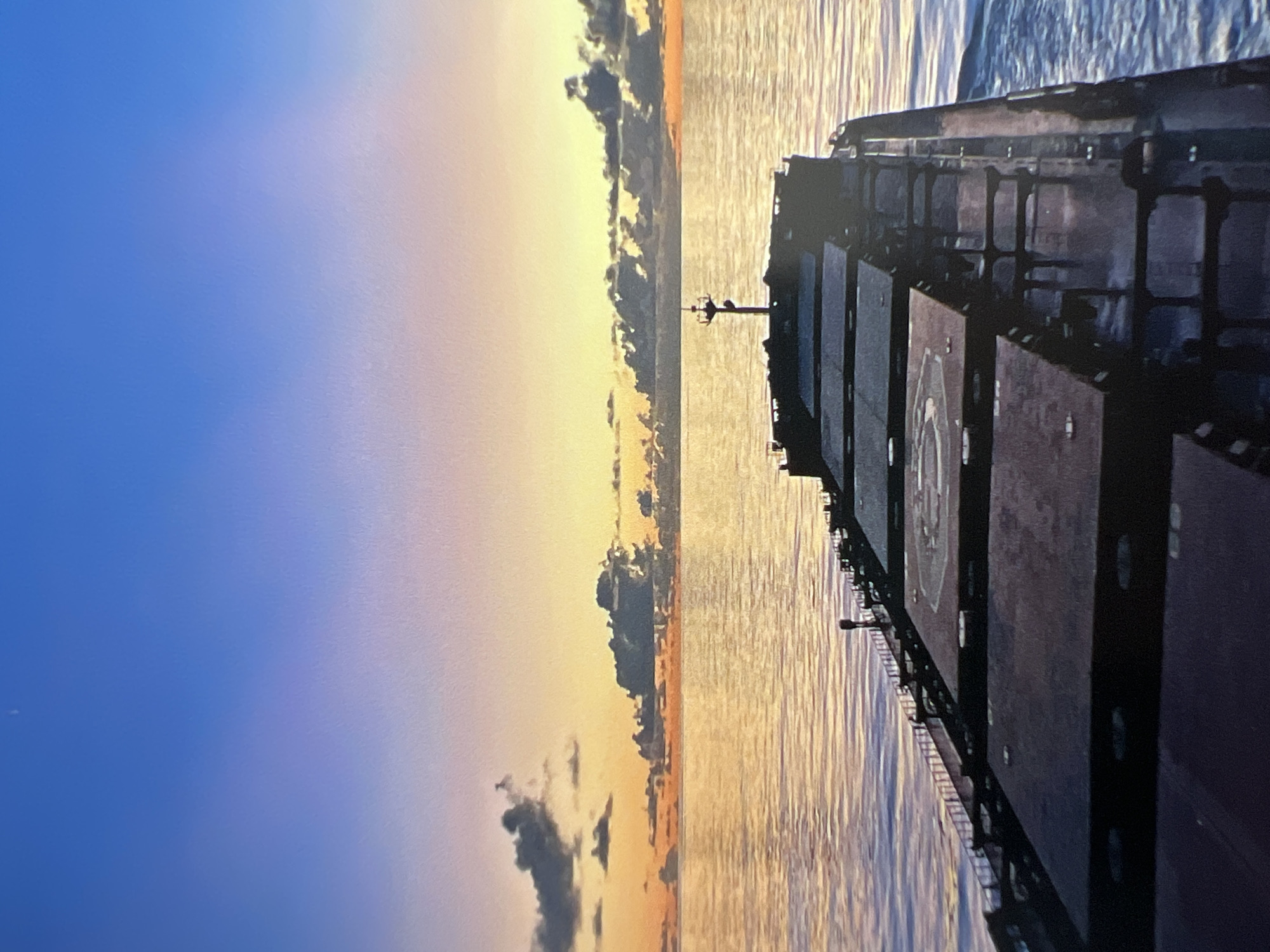 The image depicts a view from the deck of a cargo ship at sea during a sunset. The sky displays a gradient of blue and orange hues, with scattered dark clouds near the horizon. The sea reflects the warm colors of the sunset, creating a shimmering effect. The deck of the ship, which appears to be a bulk carrier, is visible in the foreground, with several cargo hatches. A helideck is marked on one of the hatches. The ship is moving, as indicated by the wake in the water. There are no people visible in the image. The overall scene is serene and atmospheric.
