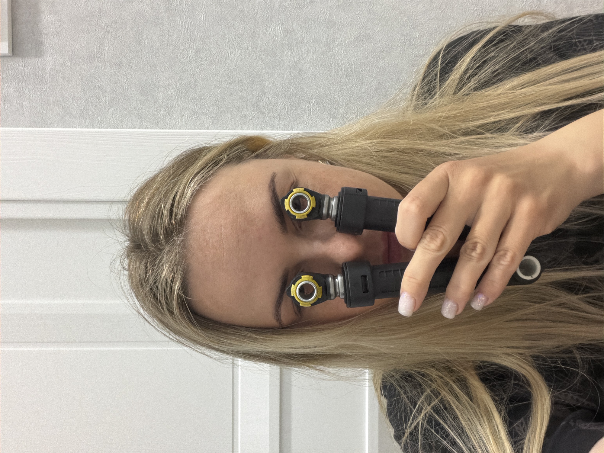 A woman with long, blonde hair is holding two black and yellow automotive components directly in front of her eyes, obscuring them from full view. The woman is facing the camera at an angle, with her head tilted to the side. Her fingernails are painted with white glitter nail polish. The background consists of a white textured wall and a white wooden panel. There are no indications of this image being taken in Votkinsk, Russia.