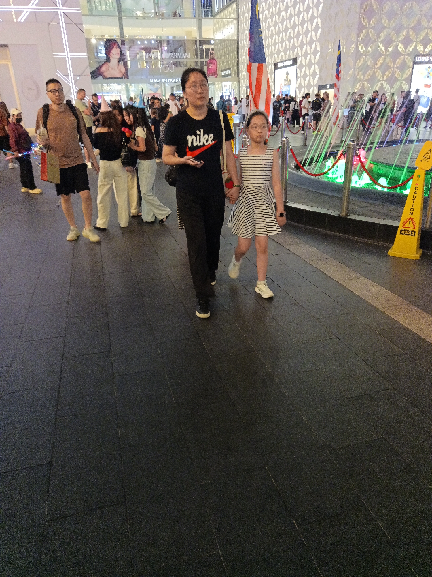 The image shows a street scene in Kuala Lumpur, Malaysia, likely in the Bukit Bintang area, known for its shopping malls and bustling atmosphere. In the foreground, a woman in a black t-shirt with a "nike" logo and a young girl in a striped dress are walking hand-in-hand. The woman wears glasses, and the girl has short hair. Behind them, a man in a brown t-shirt and black shorts is walking, carrying a bag and a drink. Further back, a group of young people, some wearing party hats, are visible, along with other pedestrians. The background features a modern building facade with illuminated displays and large flags, suggesting a commercial and entertainment district. There is also a water feature with colorful lights, and a yellow "CAUTION" sign is placed near the edge of the walkway. The ground is paved with dark tiles.