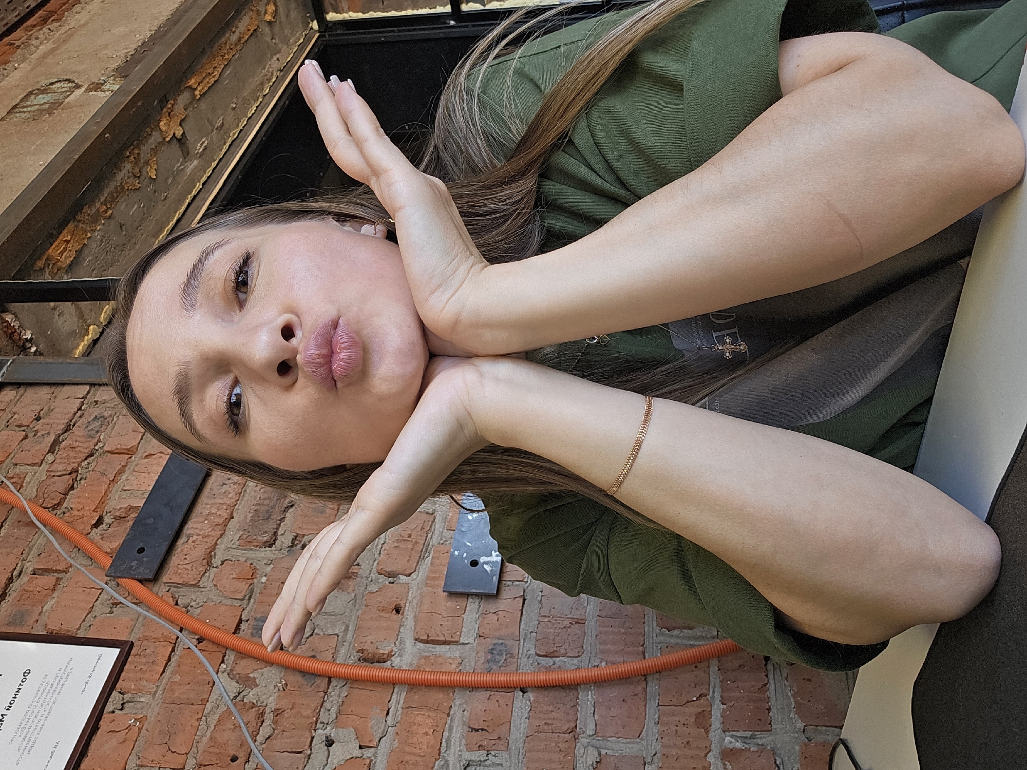 A woman with long brown hair and wearing a green shirt poses outdoors with her hands framing her face. She is making a kissing expression. The background consists of a brick wall, an orange conduit, a metal beam and a document with some text in Russian that includes "Chaikovsky".