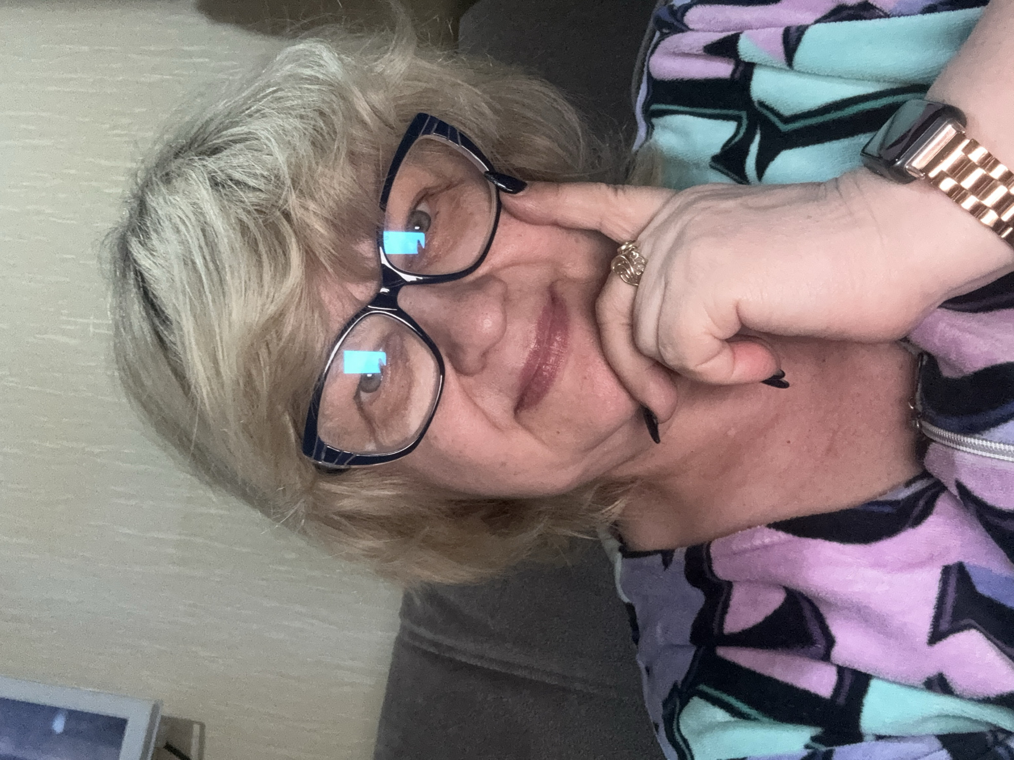 A woman with blonde hair and glasses is taking a selfie in what appears to be a living room in Mytishchi, Russia. She is wearing a colorful robe with a pattern of purple, teal, and black shapes. Her right hand is resting on her chin, and she is wearing a ring on her left ring finger and a watch on her left wrist. The background shows a textured wall and a piece of furniture.