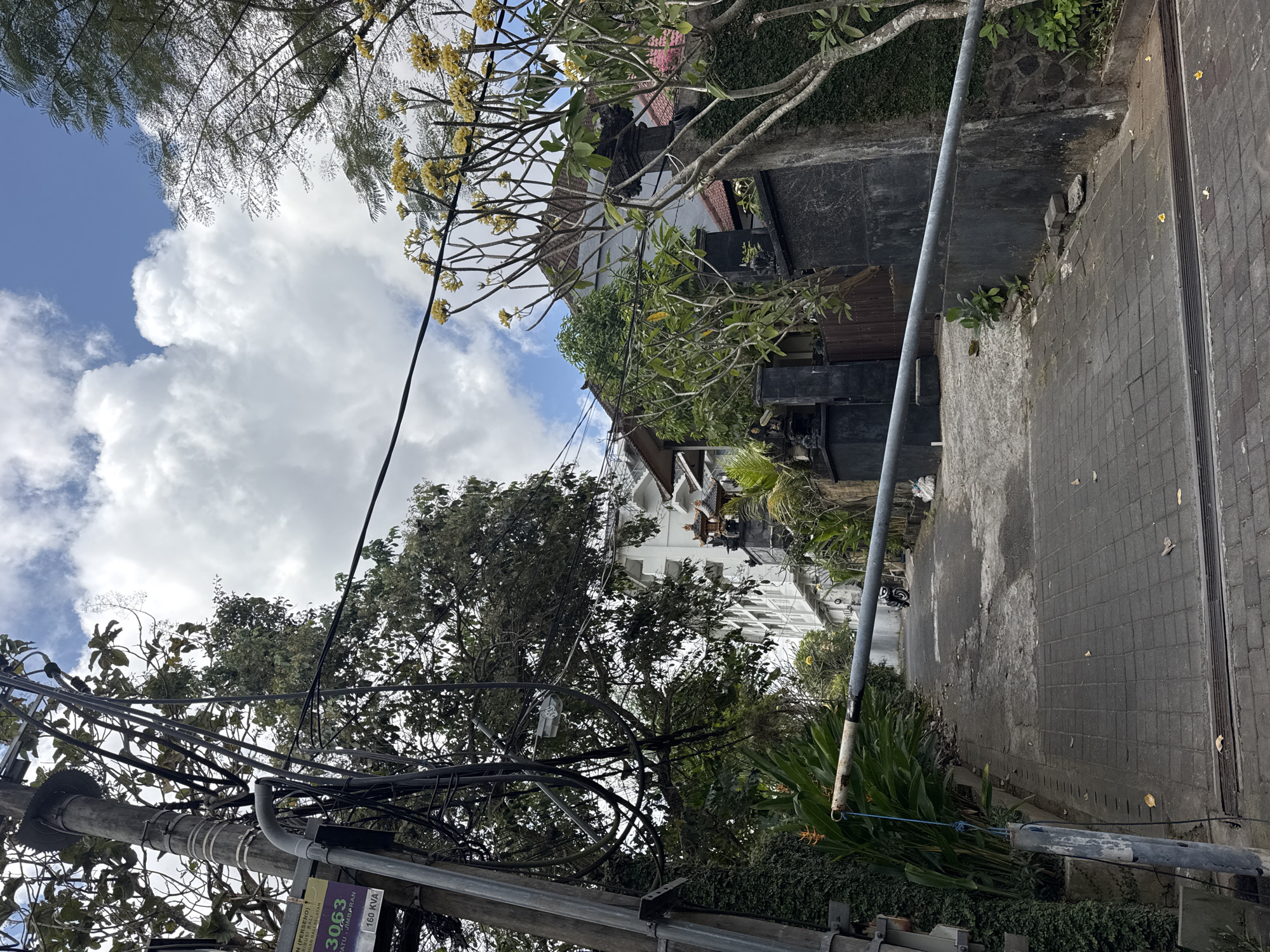 In Kuta Selatan, Indonesia, a paved pathway leads upwards, bordered by lush greenery and stone walls. A metal barrier, partially lowered, spans the path, secured by a blue rope. The pathway opens into a wider area where a motorcycle is parked. Tall trees with sparse branches and yellow flowers flank the scene. In the background, a multi-story building with white walls and tiled roofs stands against a backdrop of a bright blue sky dotted with fluffy white clouds. A utility pole with numerous black wires is prominent on the left side of the image, displaying a purple and yellow sign with the number 3063 and "160 KVA" printed on it. The overall impression is one of a tropical, somewhat cluttered, urban environment.