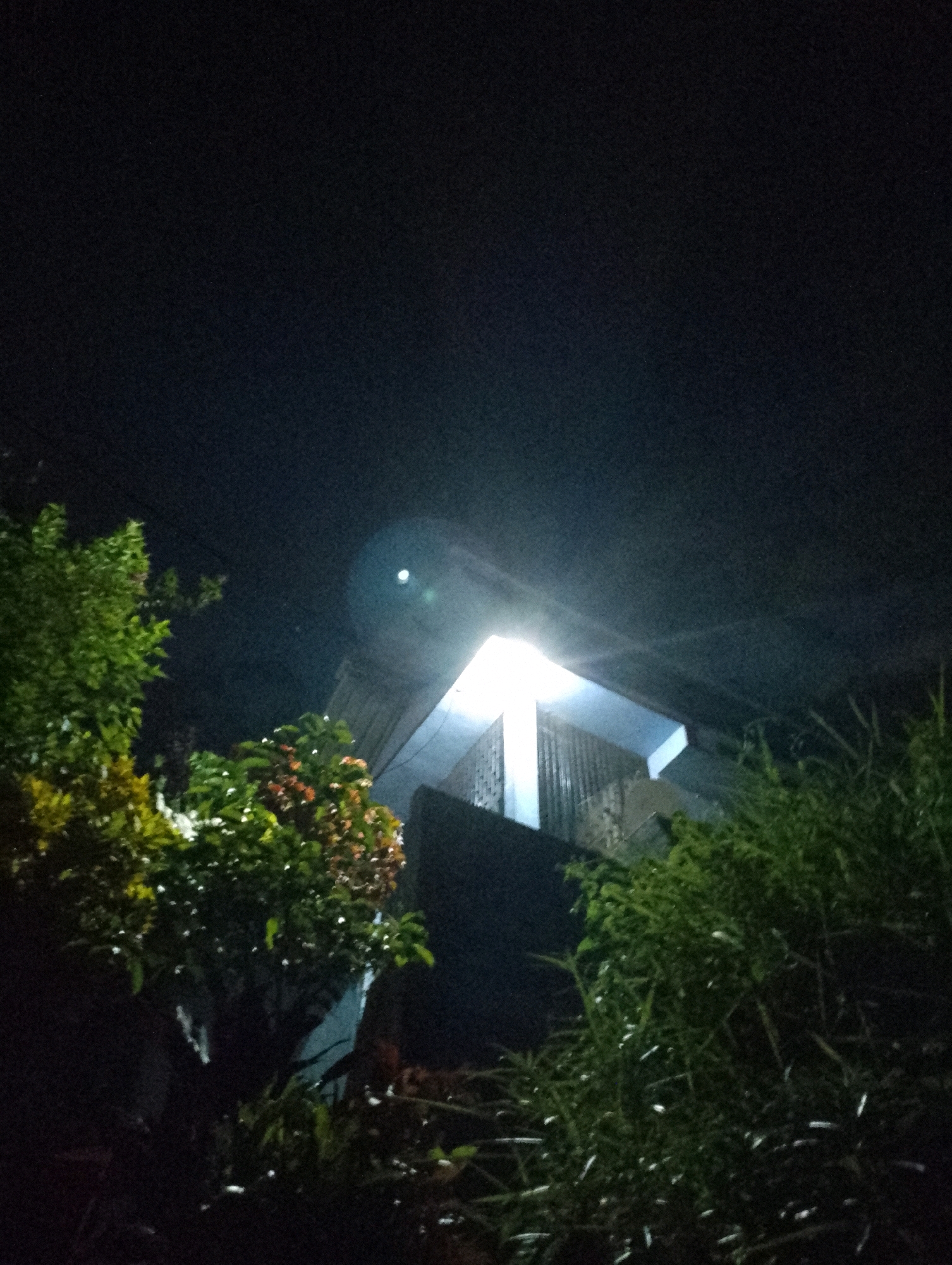 An outdoor, low-light, slightly tilted, upward-angle shot captures a building illuminated by a bright light in Maligaon, India. The building's upper section, featuring a sloped roof and what appears to be a covered balcony or walkway, is the primary illuminated subject. A strong light source, likely a floodlight, is positioned above and to the right of the frame, casting a harsh glow that washes out details in that area and creates lens flare. In the foreground and to the left, lush green foliage from trees and plants is visible, some with hints of red and yellow in their leaves. To the right and slightly behind the building, more dense green vegetation fills the lower portion of the frame. The background is a dark, night sky. No people are visible in the scene.
