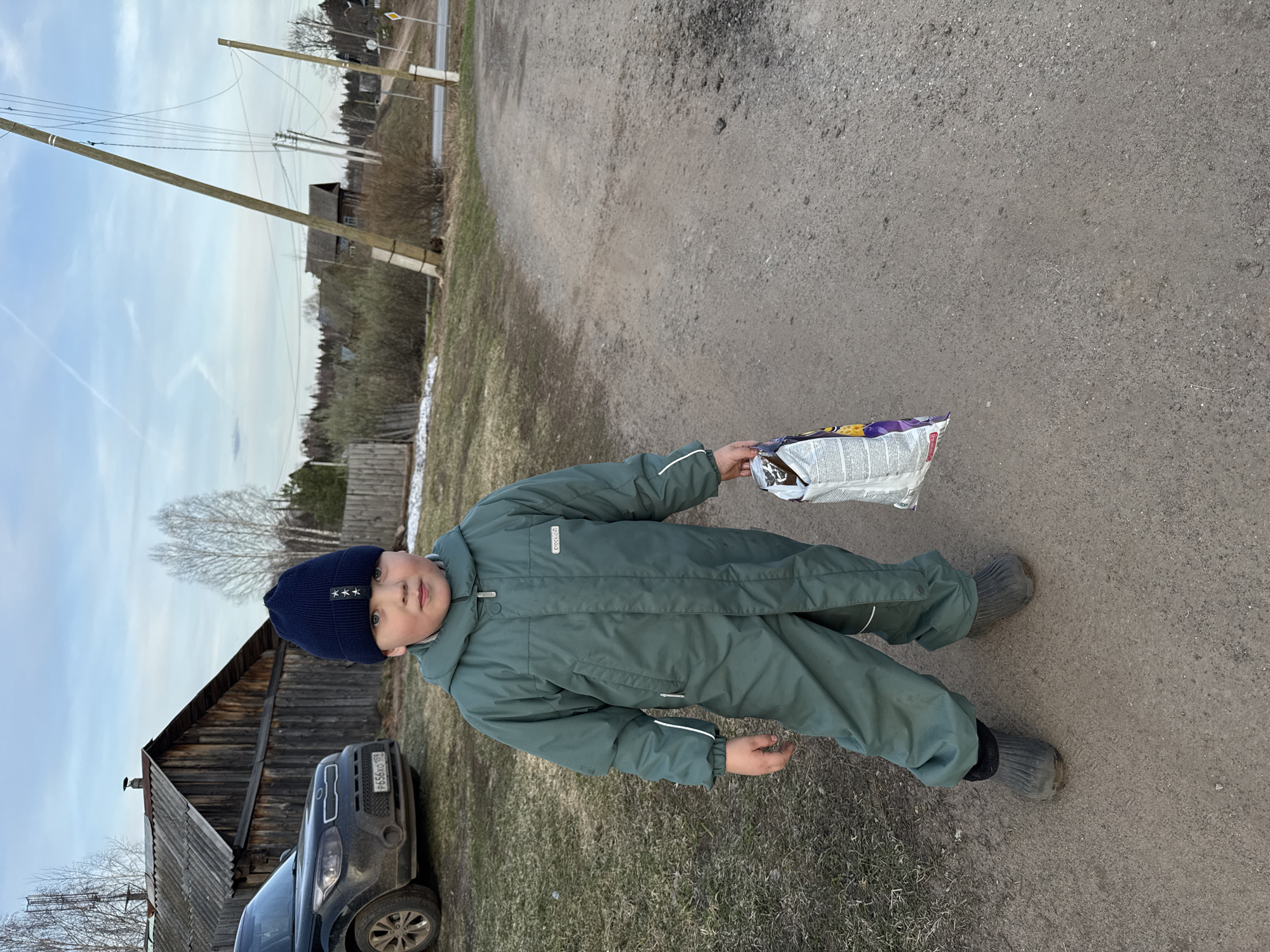 A young child dressed in a green jumpsuit and a dark blue beanie stands on a dirt road. He is holding a partially open bag of snack food in his right hand. Behind the child to the left is a dark-colored SUV parked next to a wooden building with a steeply pitched roof. The background includes a landscape with bare trees, utility poles, and other buildings. The sky is overcast. Based on the number plate, the location is likely near Zhigalki, Russia.