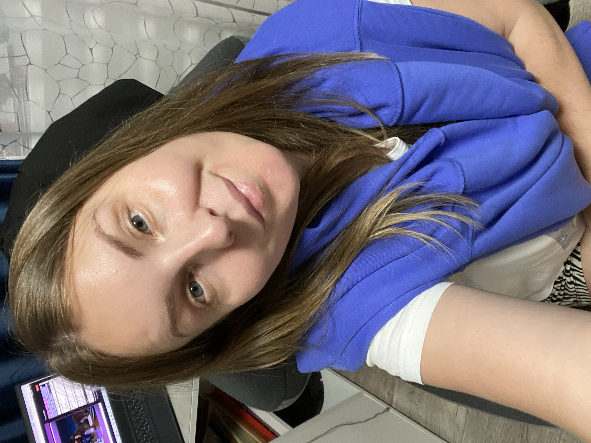 A young woman with fair skin, light brown hair, and blue eyes is shown in a close-up, selfie-style shot. She is wearing a bright blue hooded sweatshirt over a white top. A laptop is visible in the lower left corner, with a screen displaying a video or application with a purple hue. She is sitting in a black chair. The background includes a patterned wall. This image was not taken in Noviy, Russia.