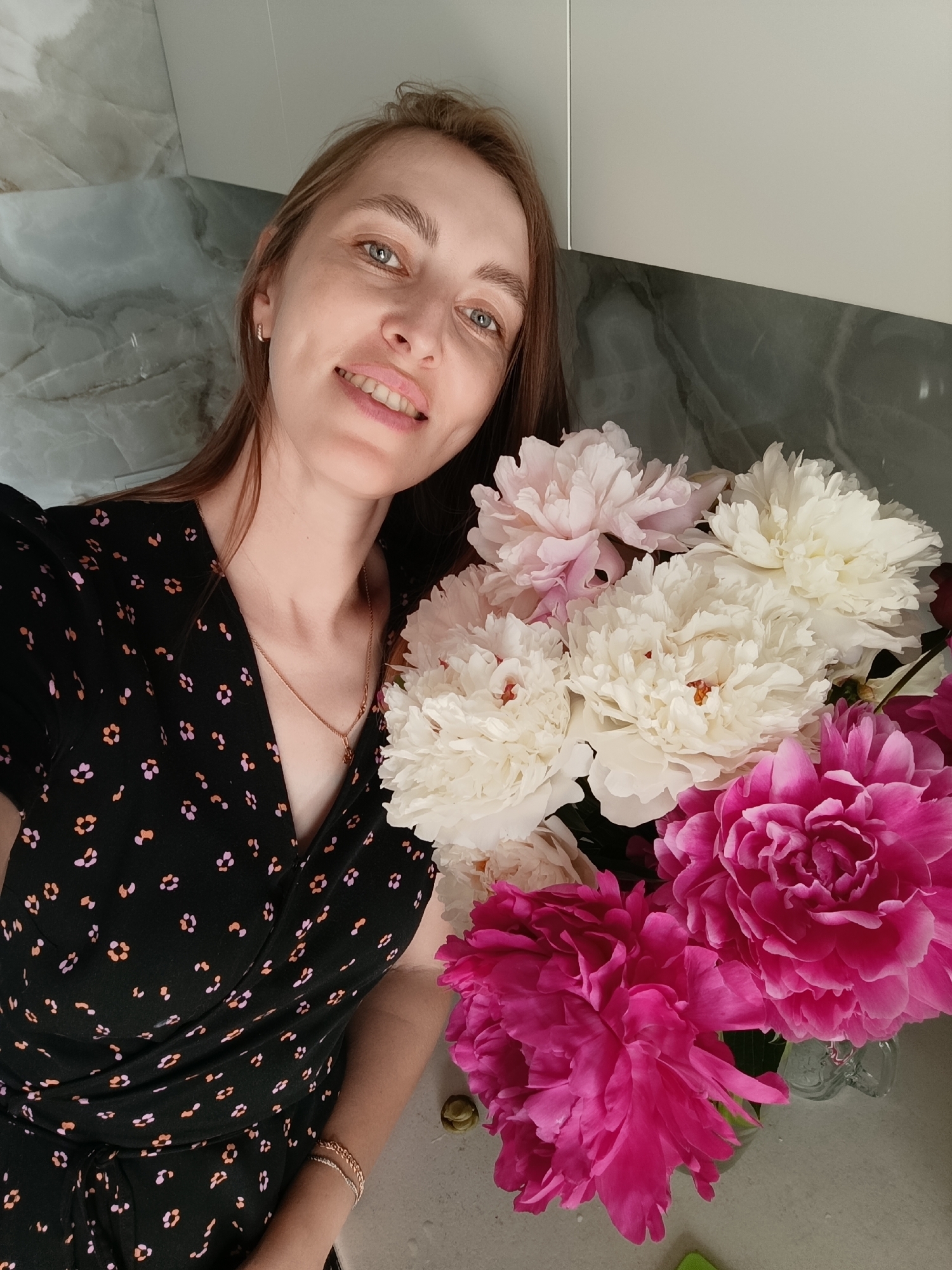A woman with long brown hair and blue eyes smiles at the camera. She is wearing a black dress with a floral pattern. In front of her is a bouquet of peonies in various shades of pink and white. The background appears to be a kitchen counter with marble-patterned backsplashes. The image was taken indoors.