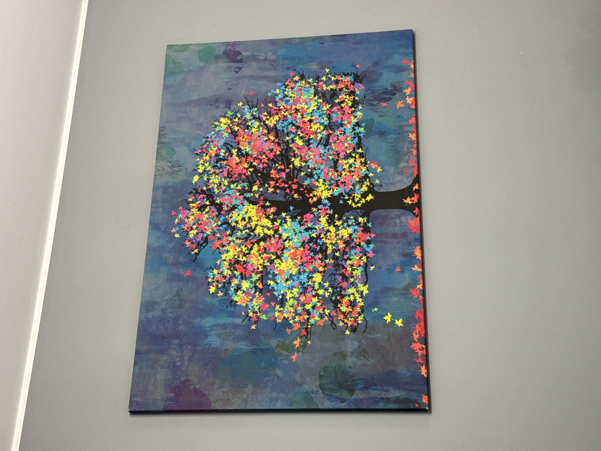 This media file shows a framed piece of artwork hanging on a grey wall. The artwork is a rectangular canvas depicting a stylized tree with a dark trunk and branches. The tree is adorned with numerous colorful, star-shaped leaves in shades of red, yellow, blue, and pink. These leaves appear to be falling, with some scattered around the tree and on the dark, moody background, which features abstract patterns in blues and purples. The style is illustrative and vibrant. The artwork is positioned at eye level on the wall, which appears to be in an indoor setting. There is a light-colored architectural element to the left of the artwork, possibly a doorway or column, also on the grey wall. No people are visible in the image. The focus is entirely on the artwork and its presentation on the wall.