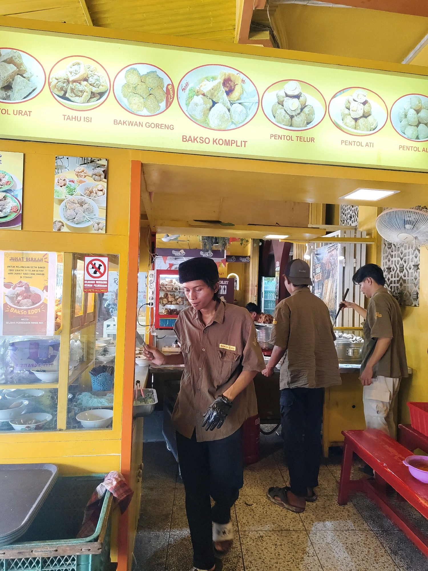 The image shows an interior scene of a food stall in Probolinggo, Indonesia. A man wearing a brown shirt, black pants, and a black cap is standing behind a counter. He is holding a knife in his right hand and appears to be preparing food. To his right, two other men in similar brown shirts are also working. One is stirring something in a pot with a ladle, while the other is standing by. The stall is decorated with yellow walls and has pictures of food items displayed on a yellow sign above. There are also posters on the glass display case in front of the counter. The environment appears to be a busy food establishment, with various serving dishes and utensils visible.