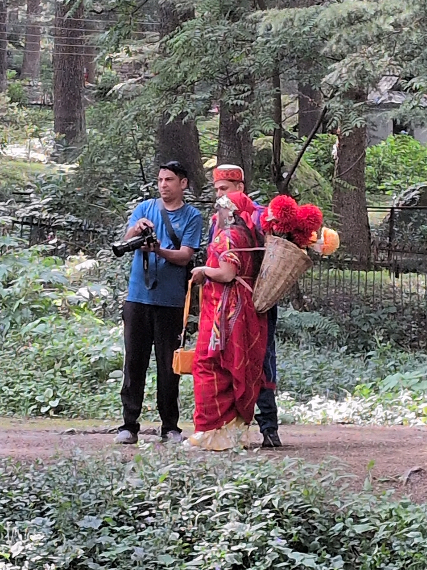 Three individuals are standing on a dirt path in a wooded area that appears to be in or near Manali, India, a hill station town in Himachal Pradesh known for its scenic beauty. The person on the left, a man wearing a blue t-shirt and black pants, holds a black camera with a strap around his neck. He is looking at the camera and seems to be taking a picture. In the center, a person in traditional attire, possibly a woman, wears a red garment with gold trim and a yellow underskirt. They have a woven basket strapped to their back, filled with red flowers and what appear to be orange marigolds. A person in the background, partially visible, is wearing a red and white patterned cap and blue pants. The surrounding environment is lush with green foliage, trees, and some natural ground cover. A metal fence is visible in the background, separating the path from denser vegetation. The overall scene suggests a moment of cultural interaction and photography amidst a natural setting.