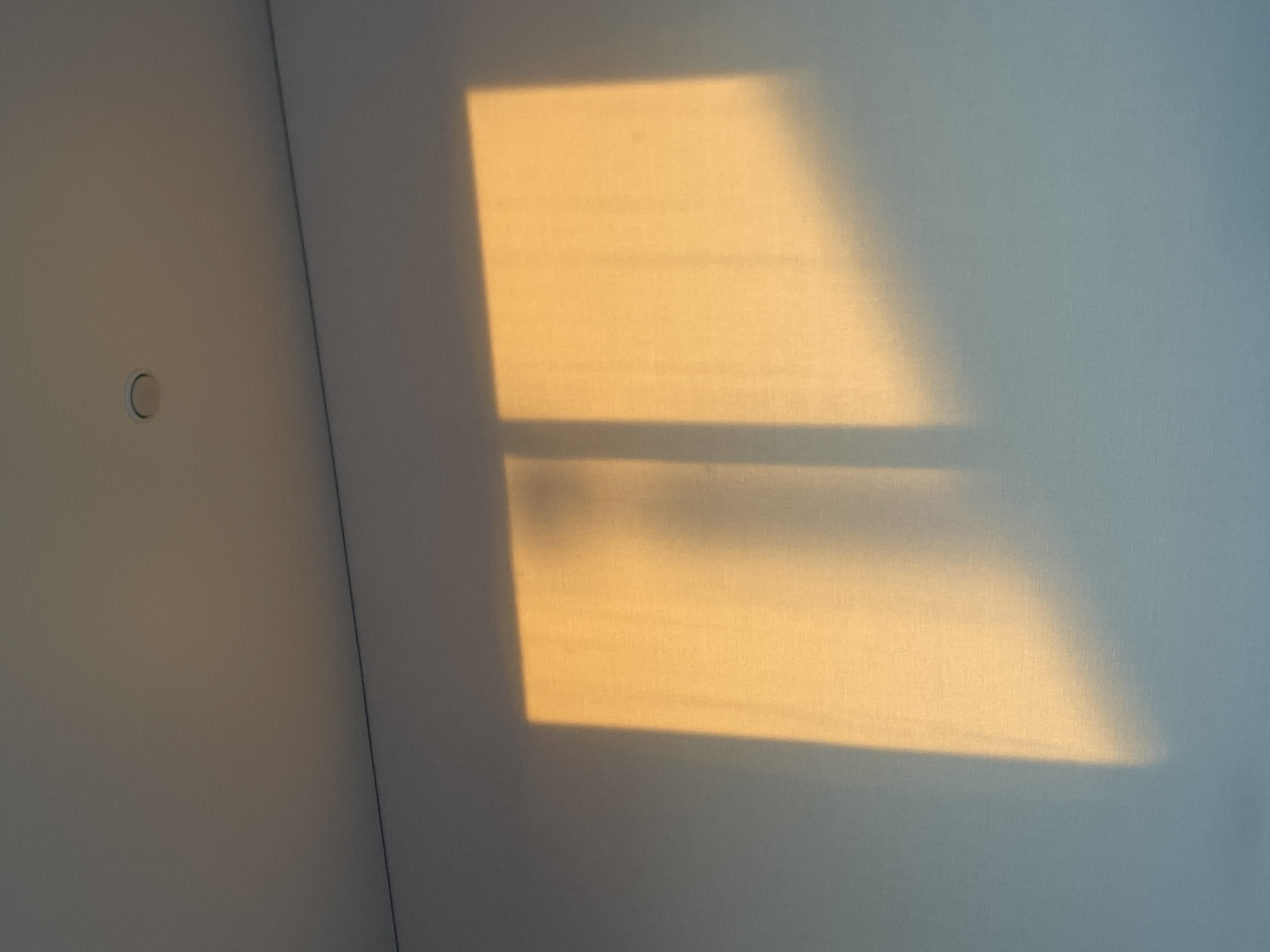 The image shows a wall in a room. Sunlight is cast onto the wall, creating a rectangular shape with a horizontal bar intersecting it. The left portion of the wall is shaded. A small, circular object is visible on the shaded wall. The texture of the wall is slightly visible. The location in the city of Подшивалово, Russia is not evident in the image.