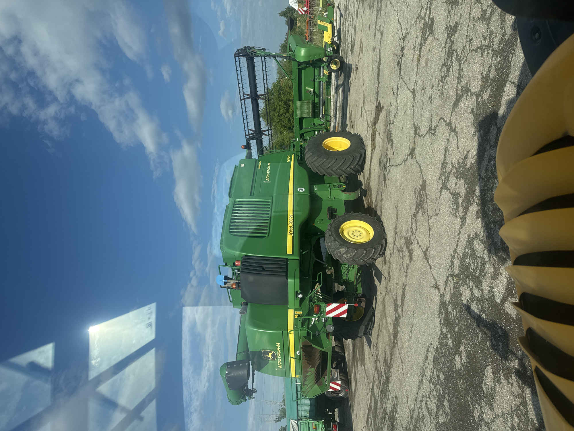 A green John Deere combine harvester with a header attached is parked on cracked asphalt under a partly cloudy sky. The text "АГРОТРОНИМ" is visible on the machine. The reflection of a window is visible in the lower left. Part of a yellow and black striped object is visible in the lower right. The location is Sarnevo, Bulgaria.