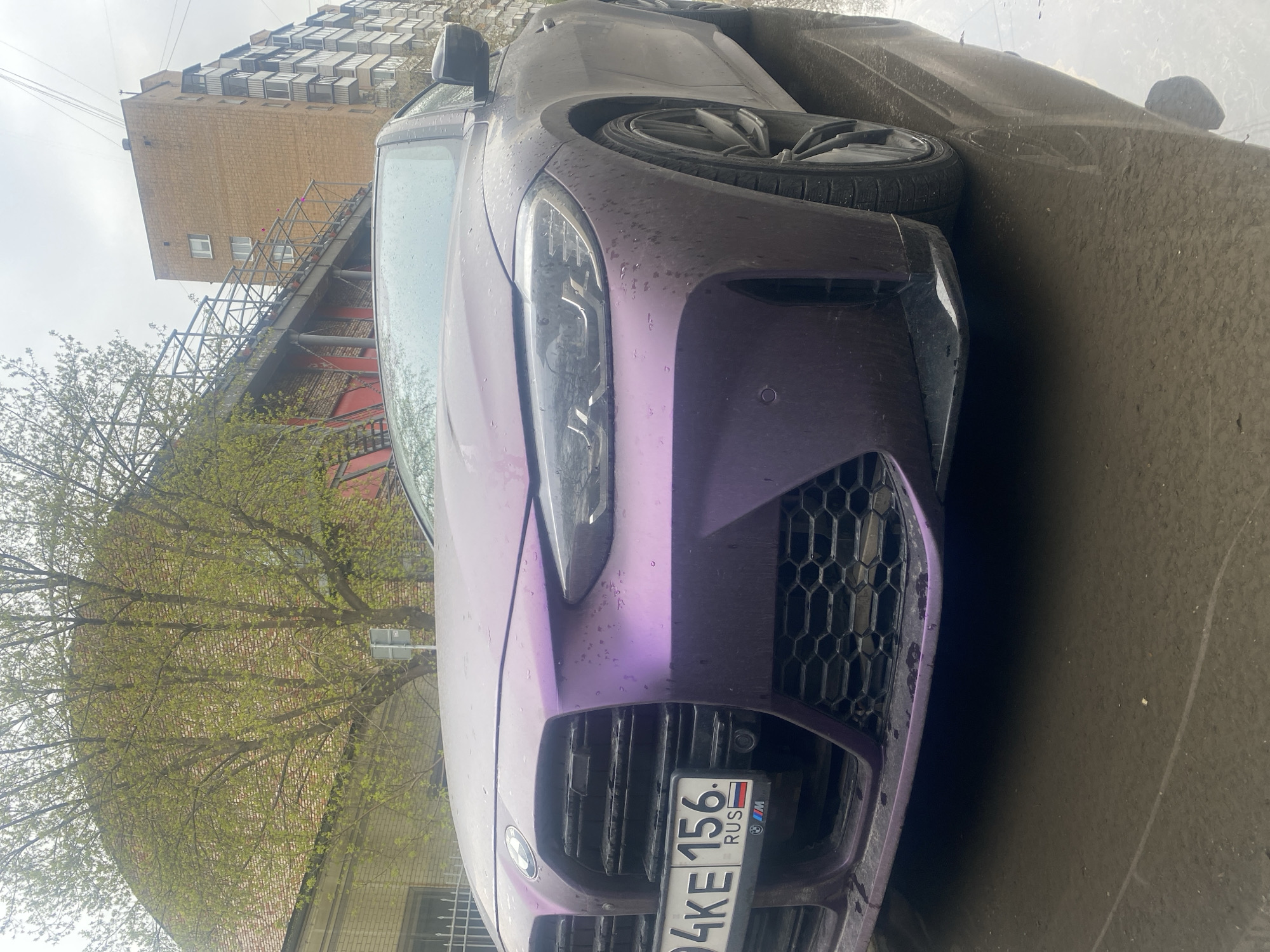 An image, captured during an overcast day, features a dirty, vibrant matte purple (or violet) modern BMW M series car, likely a G80 M3 or G82 M4, parked outdoors. The car occupies the lower and central part of the frame, positioned at an angle with its front fascia prominently displayed. Water droplets and dust cover its surface, suggesting recent rain or damp conditions.
Notable details of the car include its large, aggressive kidney grilles with a honeycomb pattern, modern LED headlights, and visible BMW logo. A Russian license plate, "94KE 156. RUS," is affixed to the front grille, with "156" indicating the Republic of Mordovia region of Russia. A small Russian flag and a BMW M logo are also present on the plate frame.
In the background, an urban environment is visible. A multi-story brick building, possibly residential or industrial, stands tall with numerous small windows. A metal support structure or walkway is attached to the side of this building. To the left, trees with fresh, light green foliage are seen, indicative of springtime. The ground appears wet, consistent with the water droplets on the car. The overall scene depicts a parked vehicle in a city setting, with no people or dynamic activity occurring. The time of day appears to be daytime, under a cloudy sky. The location context provided is Moscow, Russia, though the car's plate is registered in Mordovia.