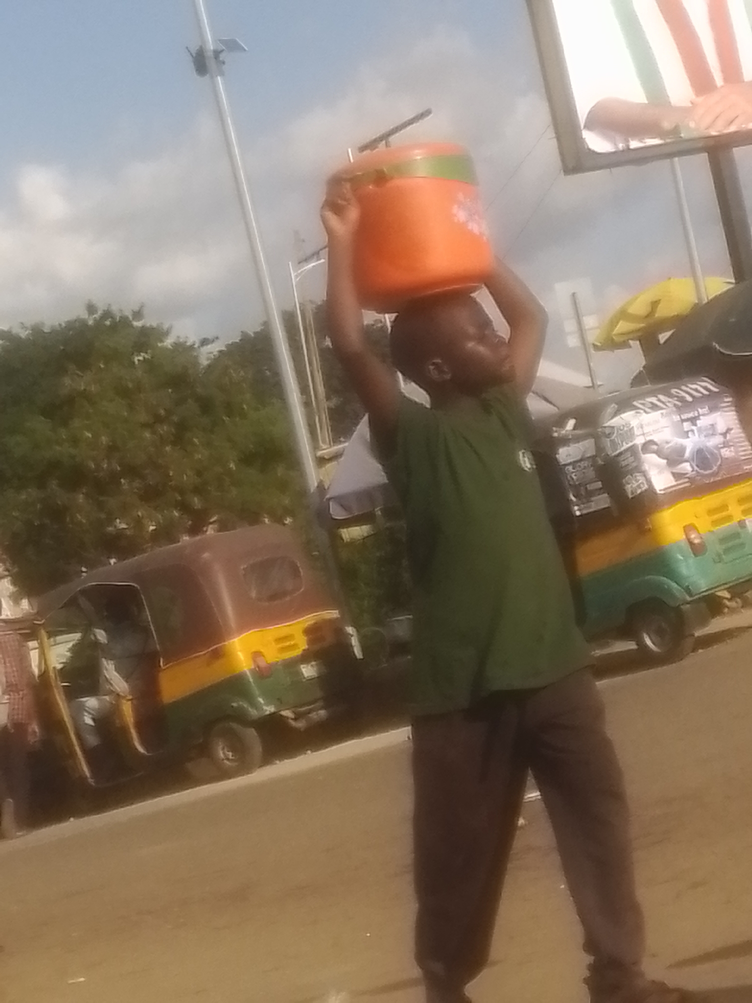 A young boy, with dark skin and wearing a green t-shirt and dark pants, carries a large, orange and green plastic container on his head. He is walking on a street, possibly in Gwong, Nigeria, as indicated by the context provided. In the background, there are two auto-rickshaws, one brown and yellow, and another with green and yellow colors. One of the auto-rickshaws has various posters and advertisements on its side. The environment appears to be outdoors, with trees and a light pole visible. The sky is cloudy and bright, suggesting daytime. The image is taken from a slightly low angle, focusing on the boy and the objects around him.