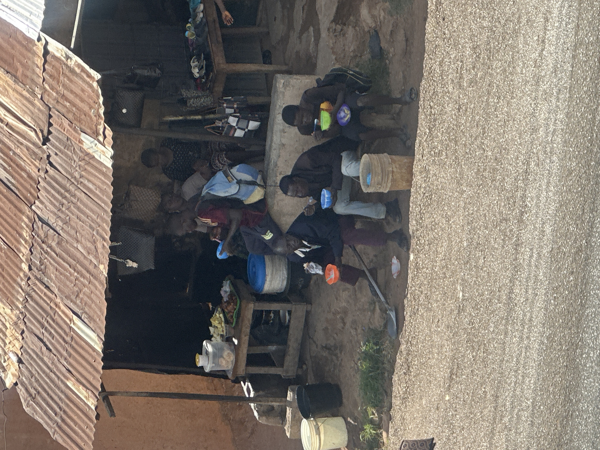 In Jos, Nigeria, a group of people are gathered outside a makeshift shelter with a corrugated tin roof. Three men are seated on the ground in the foreground, each eating from a bowl. The man on the left is wearing a dark jacket and maroon pants, holding a white piece of food in his left hand and a red bowl in his right. The middle man is wearing a dark shirt and grey pants, eating from a blue bowl. The man on the right is wearing a dark shirt and dark shorts, eating from a green bowl with a purple bowl resting in his lap. A shovel rests on the ground in front of the leftmost man. Behind them, inside the shelter, several other individuals are visible, including two women and a child. Various items are placed on a wooden table and the ground, such as buckets, containers of food, and bags. The environment appears to be an outdoor, dusty area with some sparse vegetation.