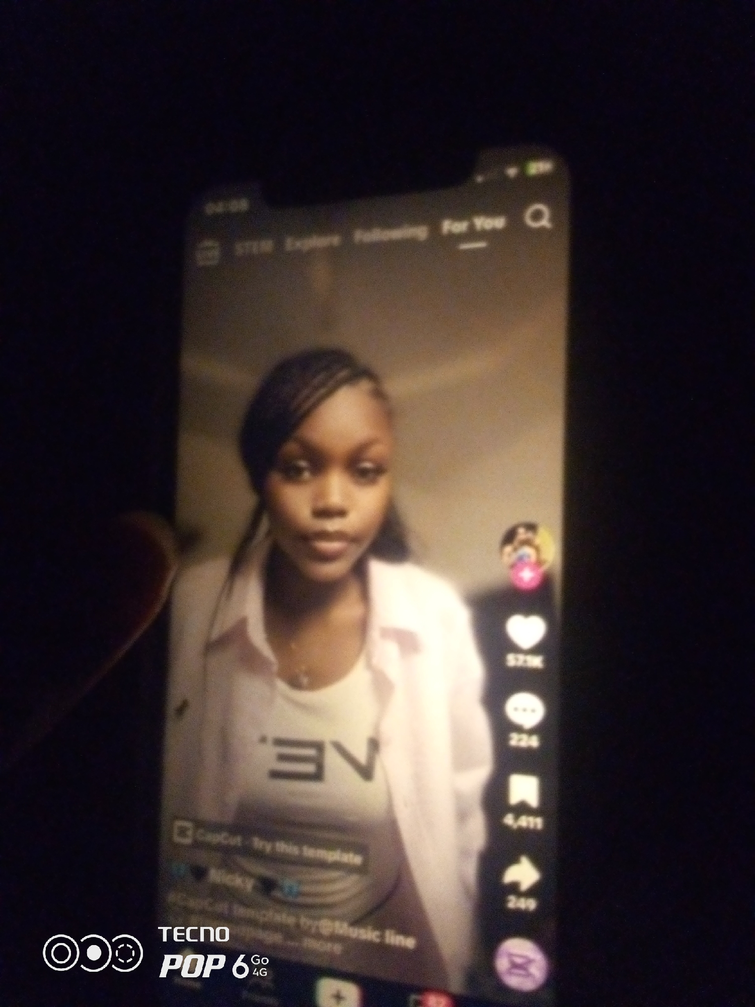 The image shows a close-up of a smartphone screen displaying a TikTok video. A young Black woman with braided hair, wearing a light pink collared shirt over a white top, is the subject of the video. Her gaze is directed towards the camera. Overlaying the video are various TikTok interface elements, including a profile icon, like count (571K), comment count (224), share count (249), and a search icon. Below the video, there are text overlays indicating "CapCut - try this template", "Nicky", and "#CapCut template by@Music line". The bottom left corner of the phone screen displays "TECNO POP 6 Go 4G". The overall environment appears to be dark, suggesting the photo was taken at night or in low light conditions, and it is not possible to determine the location in the city of Howayi, Nigeria, from the visual information provided.