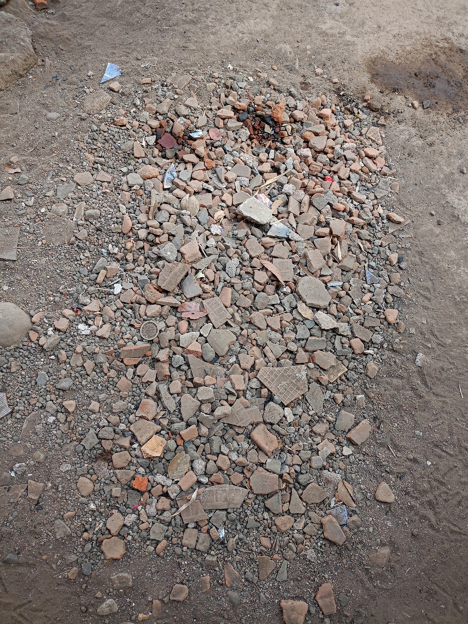 A pile of rubble, consisting of broken bricks, concrete fragments, small stones, and dirt, is situated on a dirt ground. Some shards of blue plastic are mixed in with the rubble. The scene is outdoors, with the dirt ground extending to the edges of the frame. The overall impression is one of disarray and construction or demolition debris.