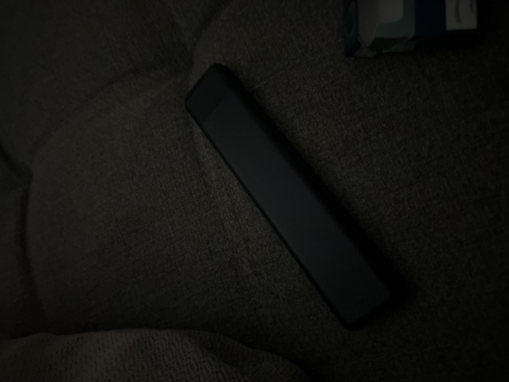 A black vape pen lies on a dark gray, textured surface, possibly a couch or fabric-covered furniture. The pen is oriented diagonally from the lower left to the upper right of the frame. To the lower right of the vape pen, a portion of a small blue and white box, possibly a cigarette pack, is visible. The lighting is dim, with some ambient light creating highlights on the vape pen and the texture of the surface. The background is dark and out of focus, suggesting a dimly lit indoor environment. No people are present in the image, and no specific location within a city is discernible.