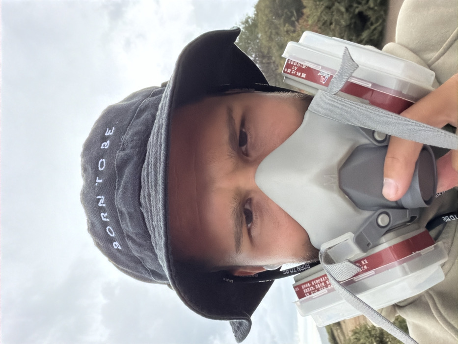 A person wearing a grey bucket hat with "BORN TO BE" vertically embroidered on it, is also wearing a grey respirator mask. The respirator has grey straps and red-colored cartridges. The individual appears to be outdoors, with trees and cloudy sky in the background. Chinese characters are visible on a label on the respirator cartridge, indicating "Plastic filter element A1".