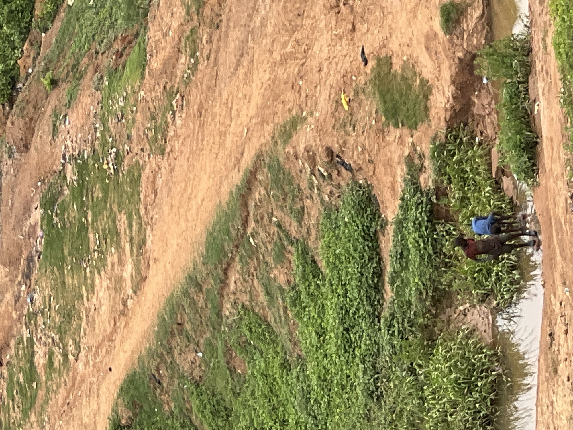 The image shows an elevated view of a landscape with a dirt path curving through it. The ground is covered with a mix of green vegetation and brown, bare earth. Two people are standing close to a body of water, possibly a stream or ditch. One person is wearing a blue shirt. The specific location is Abuja, Nigeria. The scene appears to be outdoors, with natural elements dominating the view.