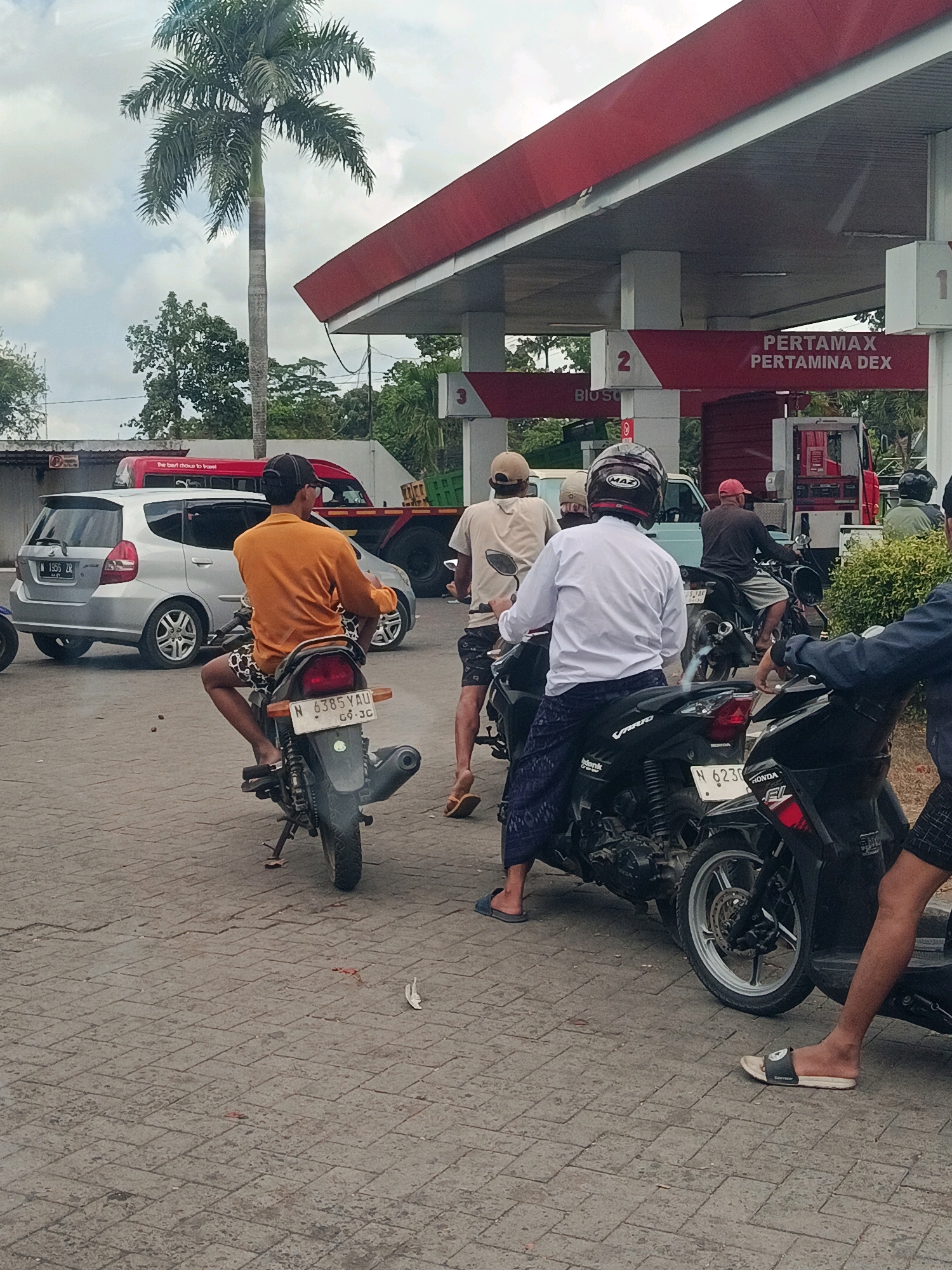 The image depicts a scene at a Pertamina gas station in Tempeh Lor, Indonesia. The foreground shows several individuals on motorcycles, appearing to be waiting or refueling. A person in an orange shirt is seated on a motorcycle with a license plate reading "N 6385 YAU 09-JC". Another person in a white long-sleeved shirt and patterned pants is on a black motorcycle. In the background, a silver car with license plate "N 1505 ZR" is visible, along with a red truck and a larger green truck. The gas station canopy, with prominent red and white signage displaying "PERTAMAX PERTAMINA DEX," is a central element of the scene. A tall palm tree stands to the left, and lush green foliage is present in the background. The ground is paved with interlocking bricks. The sky is overcast.