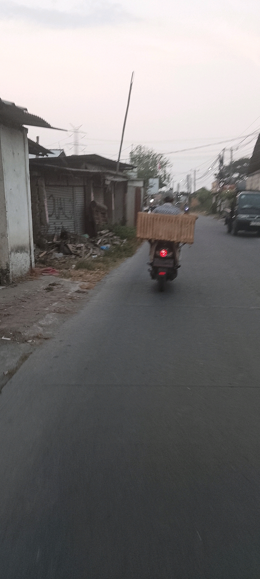 A person is riding a scooter on a street in Sukamaju, Indonesia, carrying a large, rectangular, light brown package on the back of the scooter. The scooter is dark colored with a red taillight visible. The person is wearing a blue and white checkered shirt. The package appears to be made of wood or a similar material, with a slatted construction. The road is asphalt, and there is a sidewalk or verge to the left of the road with debris and a dilapidated building with a corrugated metal roof and shuttered windows. Further down the road, other buildings and utility poles with electrical wires are visible. The sky is overcast and a light gray color. A dark colored car is visible in the background to the right of the scooter. The scene appears to be an outdoor, daytime environment.