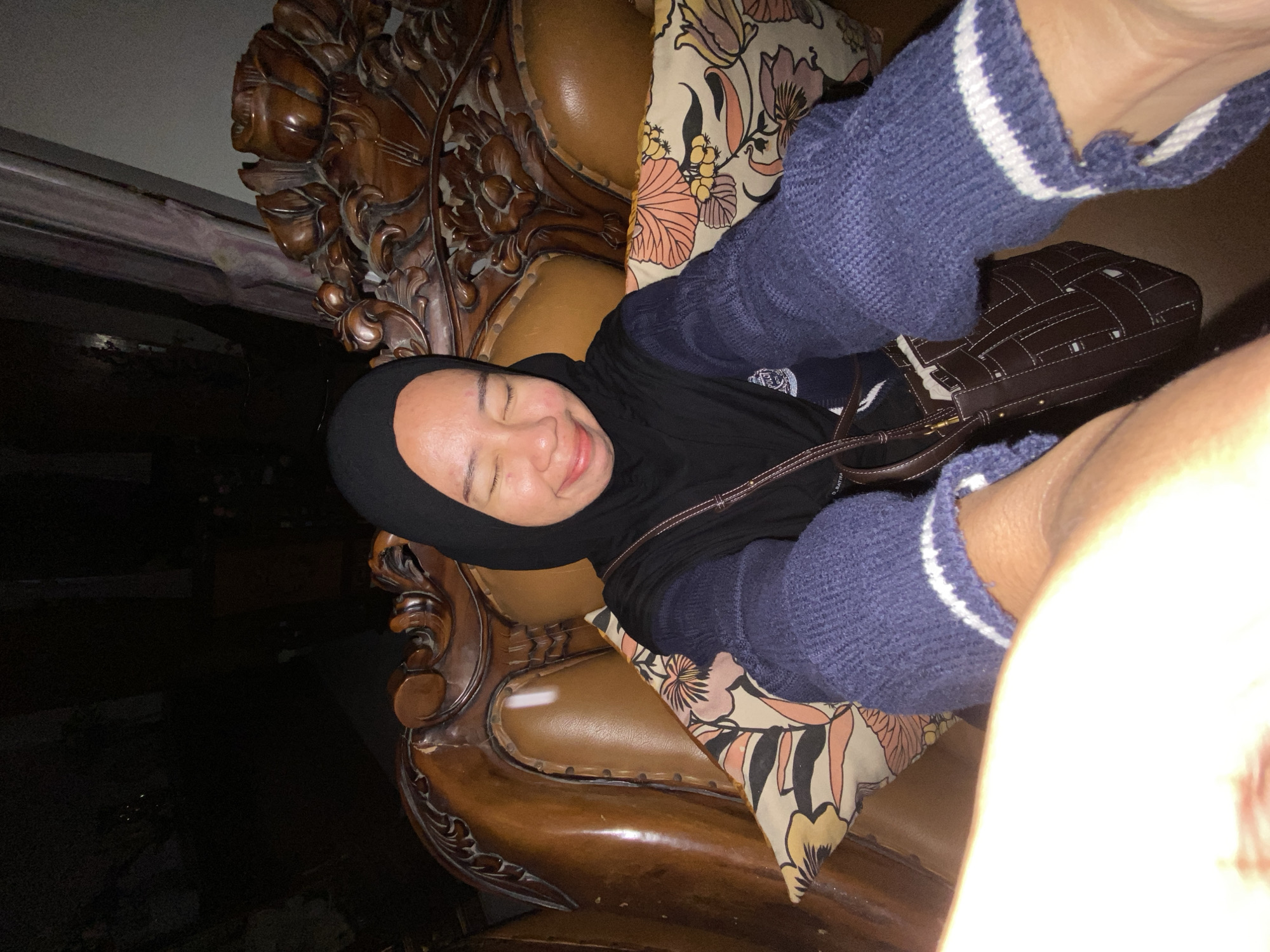 A woman wearing a black hijab and a blue sweater with white stripes on the cuffs is sitting on a wooden-carved chair in what appears to be a home in Margorejo, Indonesia. The chair has brown leather upholstery and a floral patterned cushion. The woman has her eyes closed and is smiling. She is carrying a brown woven bag. The background is dark, suggesting the image was taken indoors at night.