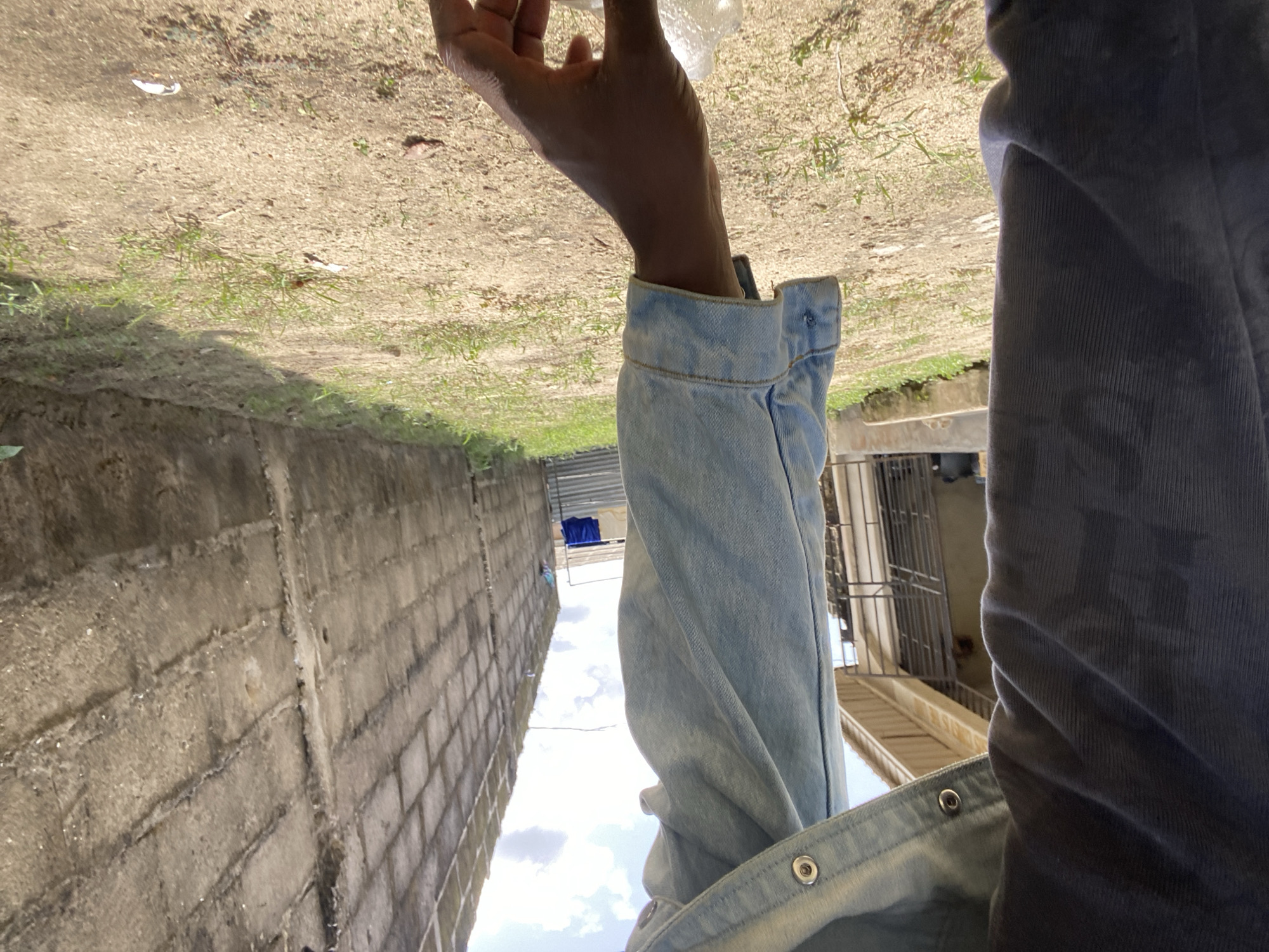 Here is a detailed description of the image:
**Scene:** The image captures a view from an individual's perspective, possibly looking down from an elevated position within a city environment.
**People:**
* An arm with dark skin tone is visible, holding a transparent object (likely a bottle or container).
* The person is wearing two jackets, one of blue jeans, and the other of dark-blue corduroy fabric.
**Objects:**
* The person is holding a transparent container.
* A brick wall is present, along with a dirt or sandy ground area with grass and vegetation.
* A building with a metal balcony or fence is visible in the background.
* Laundry is seen hanging on a wire rack in the background.
**Environment:**
* The scene appears to be outdoors with ample sunlight, as evidenced by the sky.
* The environment includes urban elements such as buildings and walls combined with natural ground features.
* The specific city remains unknown.