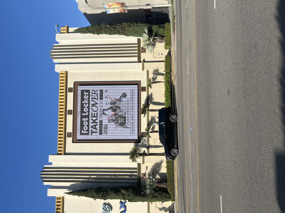 The image shows a Foot Locker store featuring a large digital billboard advertising the "Foot Locker TAKEOVER" event in Los Angeles from February 12-15 at LA Live during All-Star weekend. The advertisement highlights player appearances, exclusive drops, and fan experiences. The billboard also displays images of basketball players wearing jerseys with numbers 77 and 23. A black SUV is parked on the street next to the store.