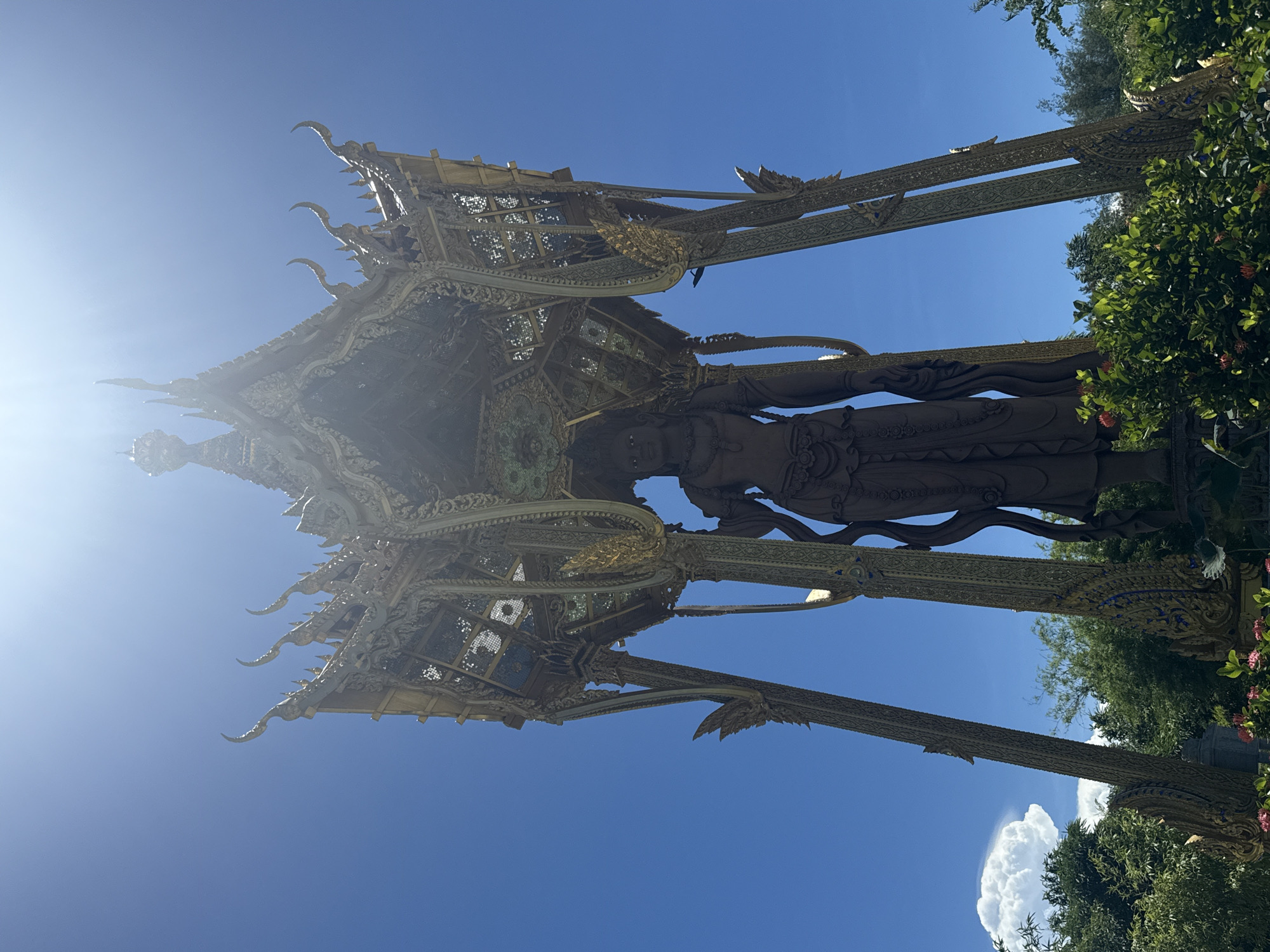 The image depicts a golden statue of a figure, likely a deity, standing beneath an ornate, open-air pavilion. The statue appears to be made of a dark, reflective metal. The pavilion has a multi-tiered roof with upward-curving eaves and elaborate carvings. The surrounding environment includes green trees and shrubs. The sky is bright blue, suggesting a sunny day. The location is Bang Pu Subdistrict Municipality, Thailand.
