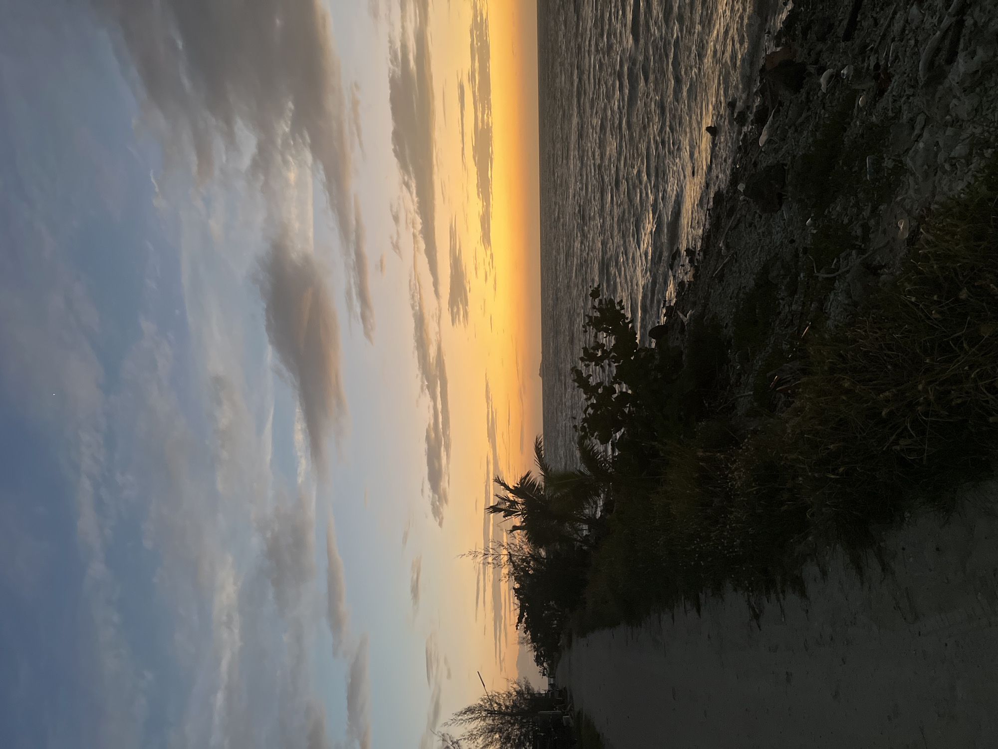 The image depicts a coastal scene at dusk, likely in the Ko Pha-ngan District of Thailand. A sandy path runs along the left side of the frame, bordered by sparse vegetation and trees, including palm trees. The path leads towards the horizon, where a calm sea meets a sky painted with hues of orange, yellow, and blue, indicative of a sunset. Scattered clouds drift across the sky. The sea is a deep, dark blue, with gentle waves lapping against a rocky shoreline. A faint light, possibly a star or a distant boat, is visible in the upper right portion of the sky. The overall atmosphere is serene and peaceful.