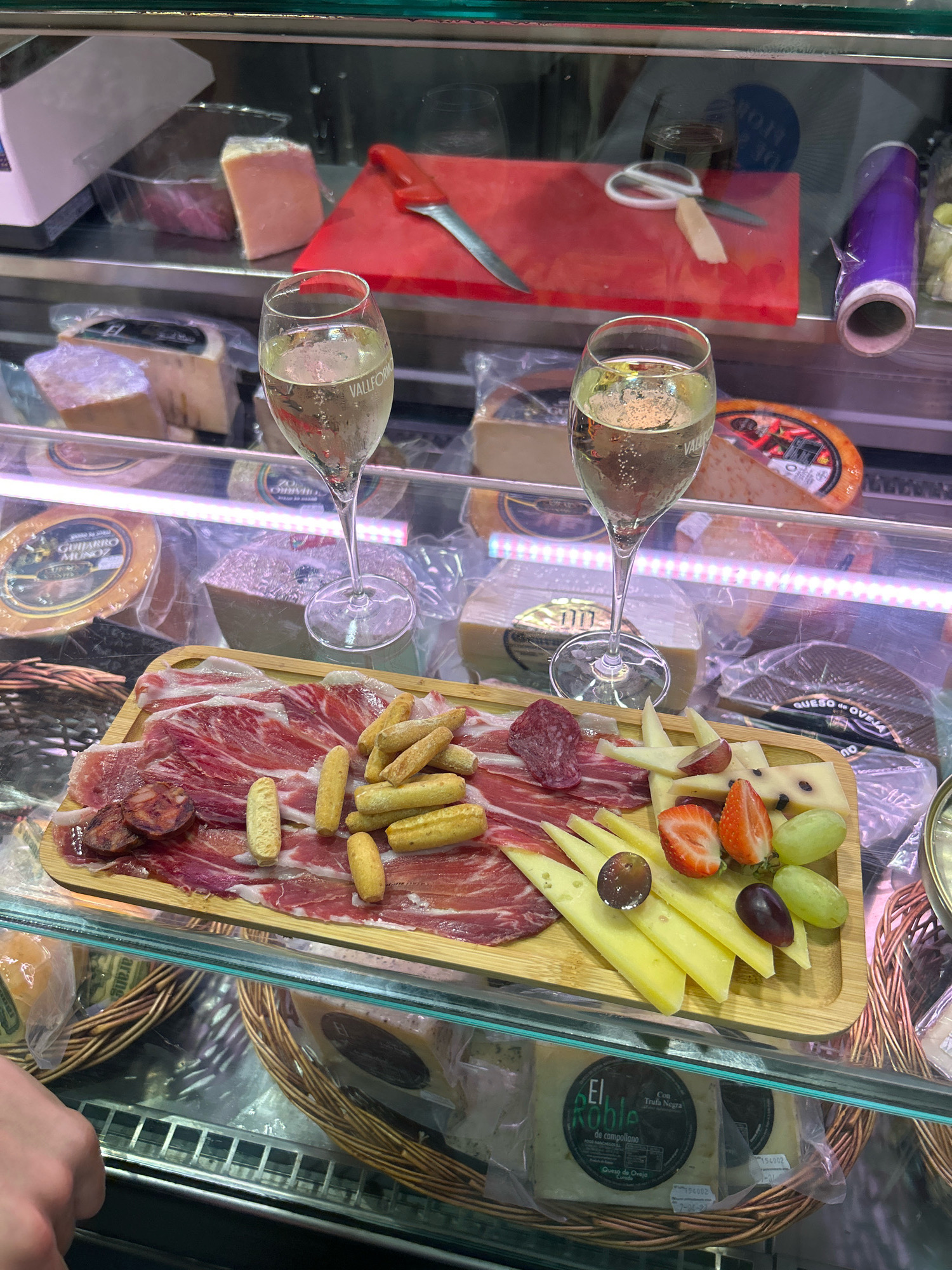 The image depicts a deli display case in Palma de Mallorca, Spain. In the foreground, a wooden board holds a charcuterie and cheese platter. The platter features thinly sliced cured ham, chorizo slices, breadsticks, slices of yellow cheese, strawberries, grapes, and a slice of dark salami. Two glasses of sparkling wine, with visible bubbles, are positioned behind the platter. The display case contains various cheeses and food products, some wrapped in plastic and others in wedges. In the background, shelves hold more food items, including wheels of cheese and a red cutting board with a knife and scissors. The overall scene suggests a food market or deli setting.