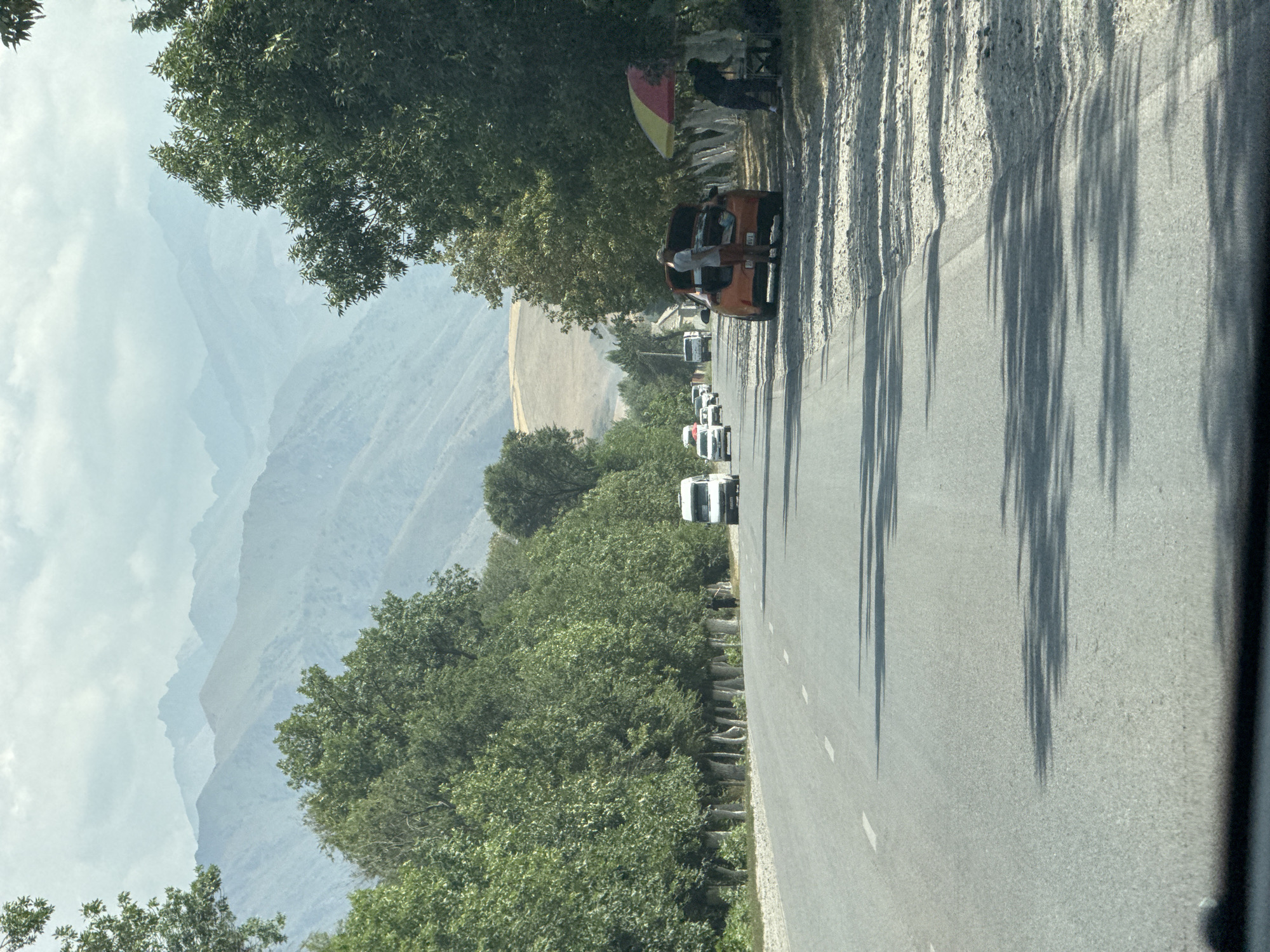 The image depicts a straight, paved road in Tash-Moinok, Kyrgyzstan, bordered by a dense line of green trees on the left and scattered trees and a roadside stand on the right. In the distance, layered, hazy mountains rise against a cloudy sky. Several vehicles are present on the road, including a white van, a few cars, and an orange sedan with its trunk open. A person stands next to the open trunk of the orange car, and two individuals are visible near the roadside stand, which has a red and yellow umbrella. The roadside to the right is covered in gravel and the shadows of trees are cast across the road, indicating it is daytime. The overall scene suggests a rural or semi-rural setting along a major thoroughfare.