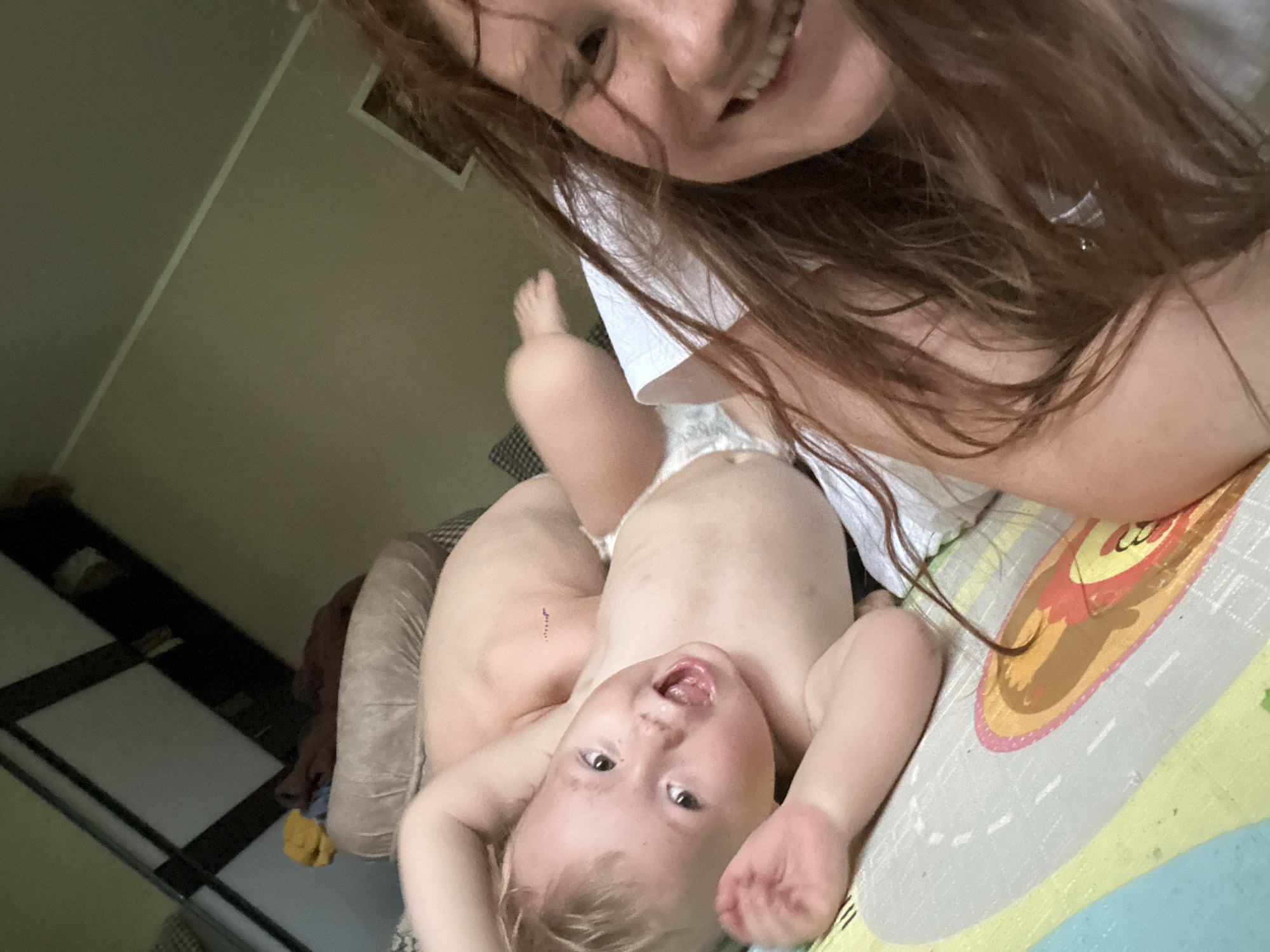 A fair-skinned woman with reddish-brown hair is holding a fair-skinned baby, likely in a diaper, lying on a colorful patterned mat. The woman is smiling and looking into the camera. The baby is lying on its back with arms raised, mouth open, and gazing toward the camera. The room has light green walls and a dark shelf or closet is visible in the background. Based on the context, it is unknown if the location is in Nizhny Novgorod, Russia.